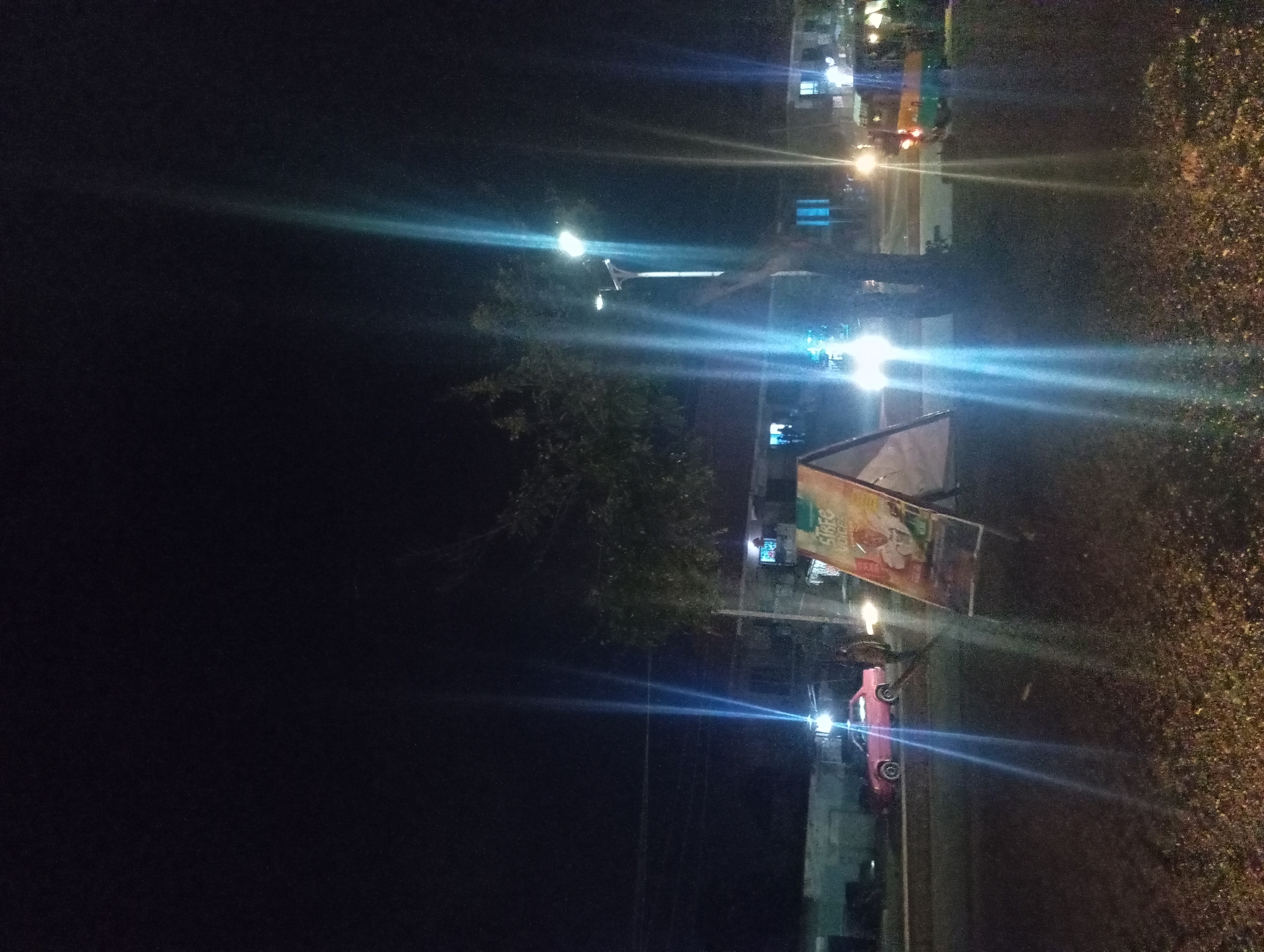 The image depicts a nighttime street scene with several vehicles and buildings illuminated by artificial lights. A red car is visible on the right side of the image, facing left. In the background, there are tall buildings with numerous windows, suggesting an urban environment. Streetlights and the lights from the buildings create bright streaks of light across the dark sky, indicating a long exposure photograph or lens flare.
A billboard with text in an unknown language is mounted on a pole. Trees with dense foliage are present, some appearing to be illuminated by the surrounding lights. The overall impression is of a bustling street at night, possibly in a commercial or residential area. The lighting conditions suggest it is dark, with no natural light sources visible. There are no people clearly identifiable in the image. The scene is static, with no specific activity or event being captured beyond the presence of vehicles and stationary structures. The image appears to be taken from an elevated position, looking down onto the street.
