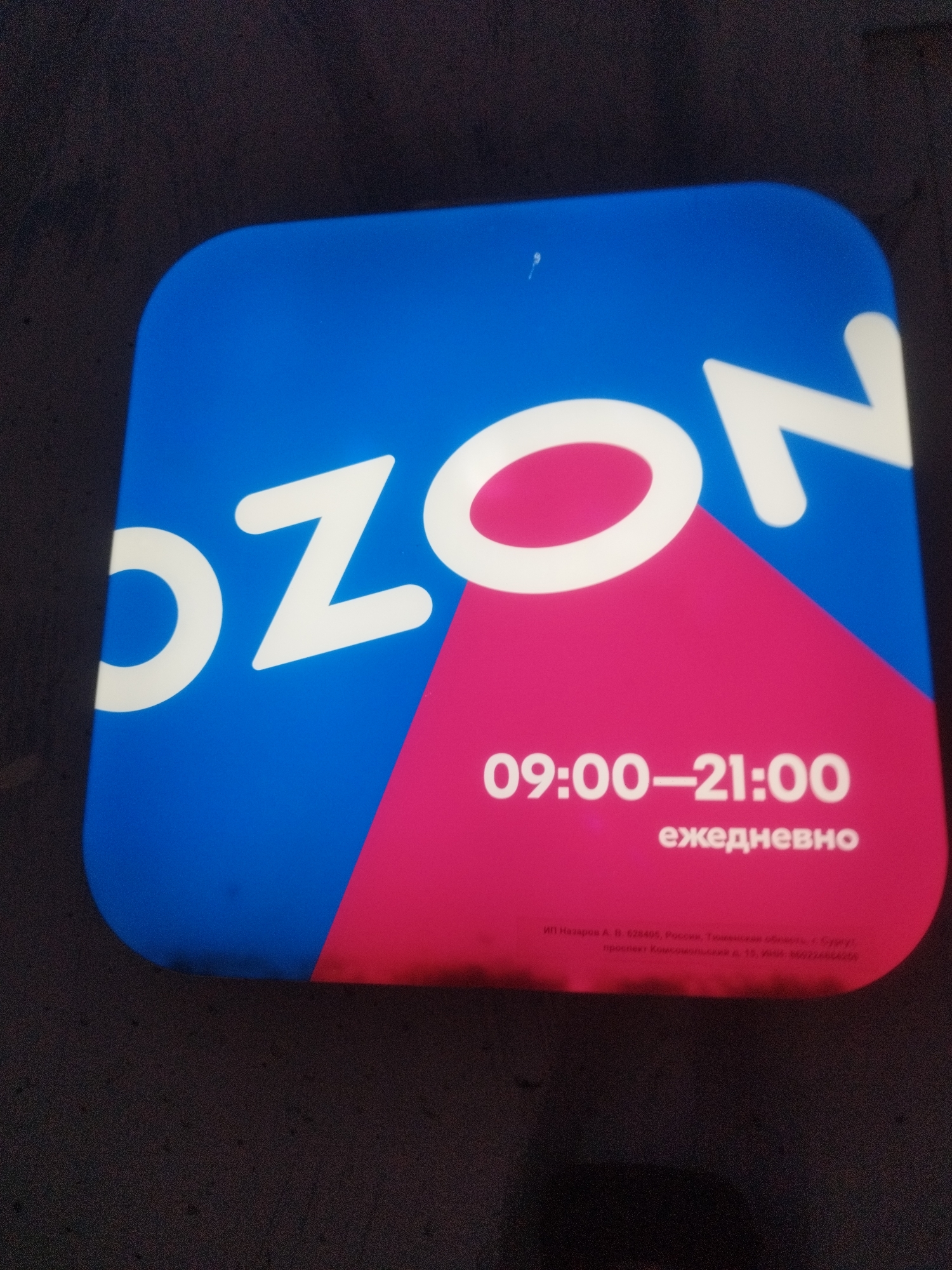 A digital sign displays the Ozon logo, which consists of the letters "OZON" in white against a blue background. A pink triangular shape is superimposed on the lower right portion of the blue background. Below the logo, the text "09:00-21:00" and "ежедневно" (daily) are displayed in white. At the bottom of the sign, there is small text, possibly an address or legal information. The sign is illuminated and appears to be mounted outdoors or in a public space. The background of the image is dark and textured, suggesting it is likely nighttime or an indoor setting with low lighting. There is no indication of the specific location in Surgut, Russia, or any people present in the image.