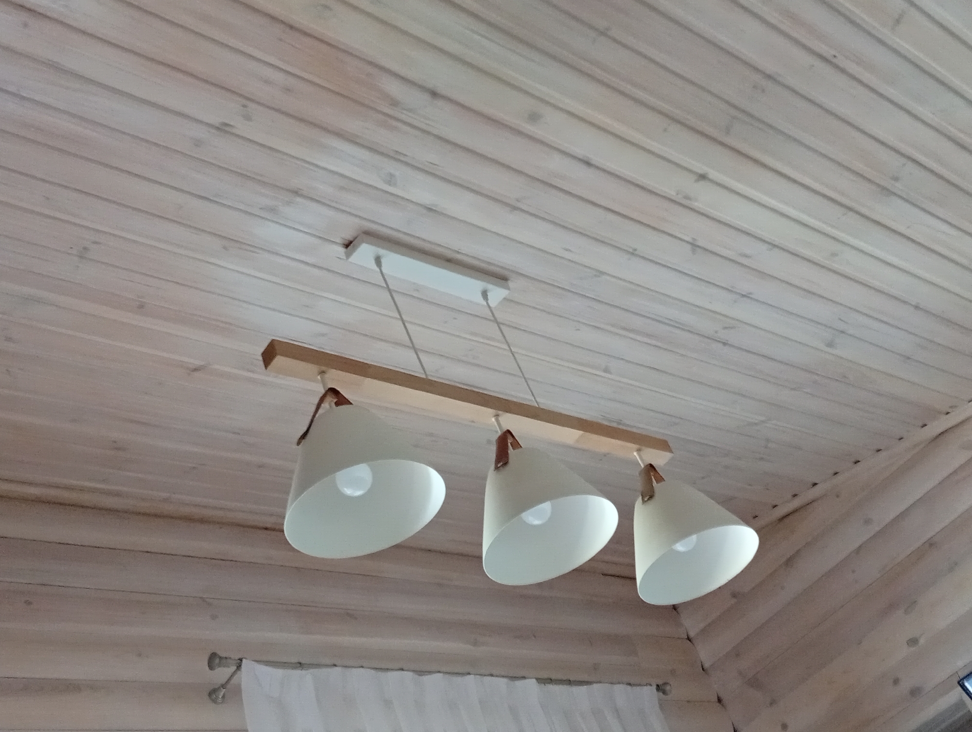 The image shows a wooden ceiling with a pendant light fixture. The fixture has a light-colored rectangular mounting plate on the ceiling from which three white conical lampshades hang. Each lampshade has a brown leather strap that attaches it to the fixture. The lampshades are arranged in a line and appear to be made of metal. The ceiling is constructed of light-colored wooden planks. The scene is viewed from below, looking up at the ceiling and the light fixture. There are no people or animals visible in the image. The location is identified as Sokyrna, Ukraine.