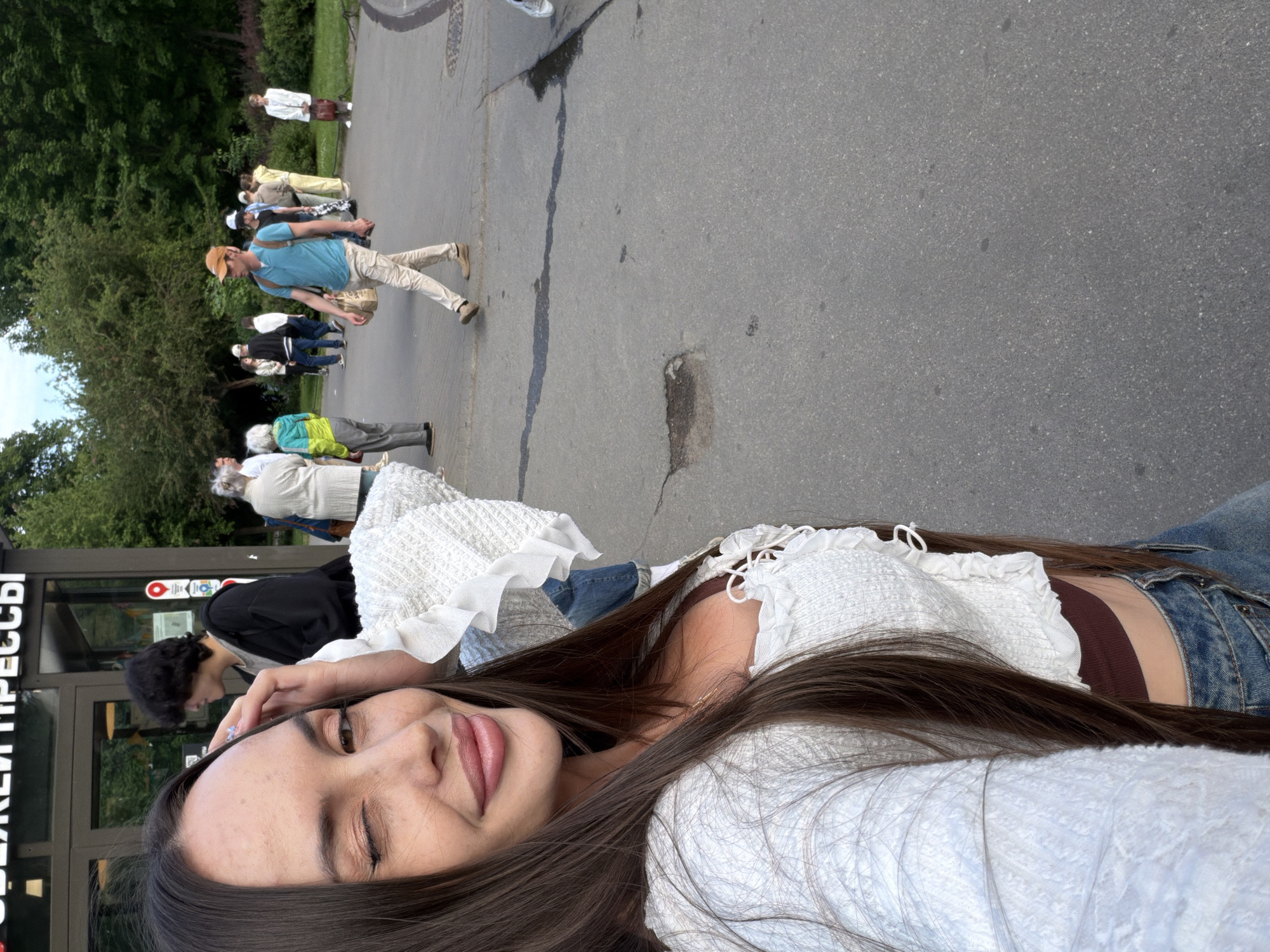 The image shows a woman taking a selfie in front of a kiosk in Saint Petersburg, Russia. The kiosk appears to be a food or drink stand, with text in Russian visible on its facade. Behind the woman, a line of people is waiting, some of whom appear to be tourists. The background features a park-like setting with trees and green foliage. The woman is wearing a white ruffled top and jeans, and she is winking at the camera. The overall atmosphere suggests a casual outing in a public space.