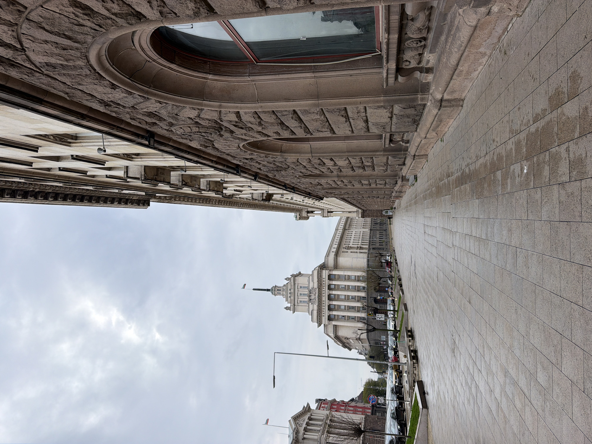 The image captures an urban street scene in Sofia, Bulgaria, on an overcast day. The perspective looks down a wide, paved thoroughfare or plaza.
In the foreground, to the left, a grand building with a rough-hewn stone facade features an ornate, arched window near its upper section, exhibiting classical architectural elements. The paved area, which appears wet in patches suggesting recent rain, extends towards the background.
The dominant feature in the midground and background is the iconic National Assembly of Bulgaria (Narodno Sabranie). This large, light-colored neoclassical building stands prominently with its distinctive columns and a central spire. A Bulgarian flag flies from a flagpole atop the spire. The building's facade clearly displays the text "НАРОДНО СЪБРАНИЕ" (National Assembly) in Cyrillic script.
To the right of the paved area, a street runs parallel, lined with parked cars, lampposts, and some bare trees, indicating perhaps a late autumn or winter setting. Another building further down the street also displays a Bulgarian flag. The sky is predominantly cloudy, contributing to the muted lighting of the scene. No people are visible, lending a quiet atmosphere to the otherwise bustling city center.