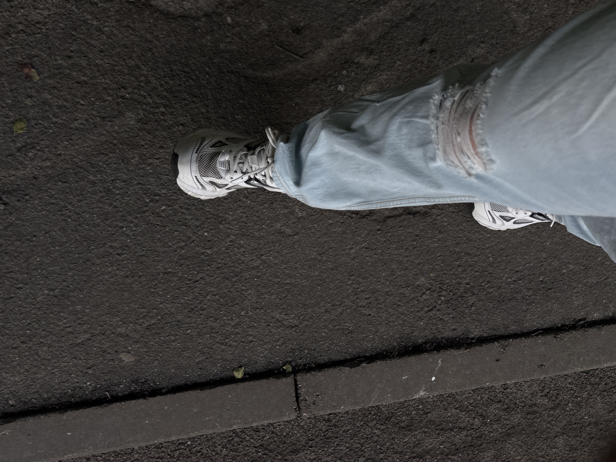 The image shows a close-up view of a person's legs and feet wearing light-wash ripped jeans and white sneakers. The person is walking on an asphalt surface next to a curb. The scene is outdoors, and the lighting suggests it is daytime. The ground is dark and textured, with some small debris and a few green leaves visible. A narrow, raised concrete curb separates the asphalt from a darker, rougher surface. One of the jeans has a prominent rip on the knee, revealing skin underneath. The sneakers are athletic in style, with a mesh upper and detailed sole. The image is taken from a slightly elevated angle, looking down. There is no visible text in the image. The overall impression is casual and urban.