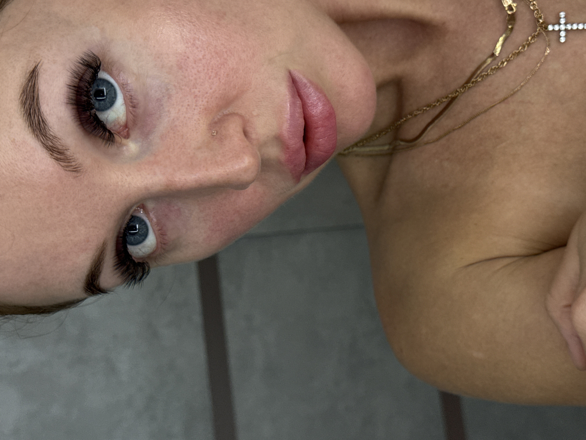 The image is a close-up, angled shot of a woman's face and upper torso. Her face is turned towards the viewer, showing her blue eyes with long, dark eyelashes. Her eyebrows are well-defined. She has a nose piercing on the left side. Her lips are pink and appear to have lip gloss on them. The woman is wearing a gold necklace with a cross pendant. Part of her bare shoulder and upper chest are visible. The background is a neutral gray color. The city is not evident in this image.