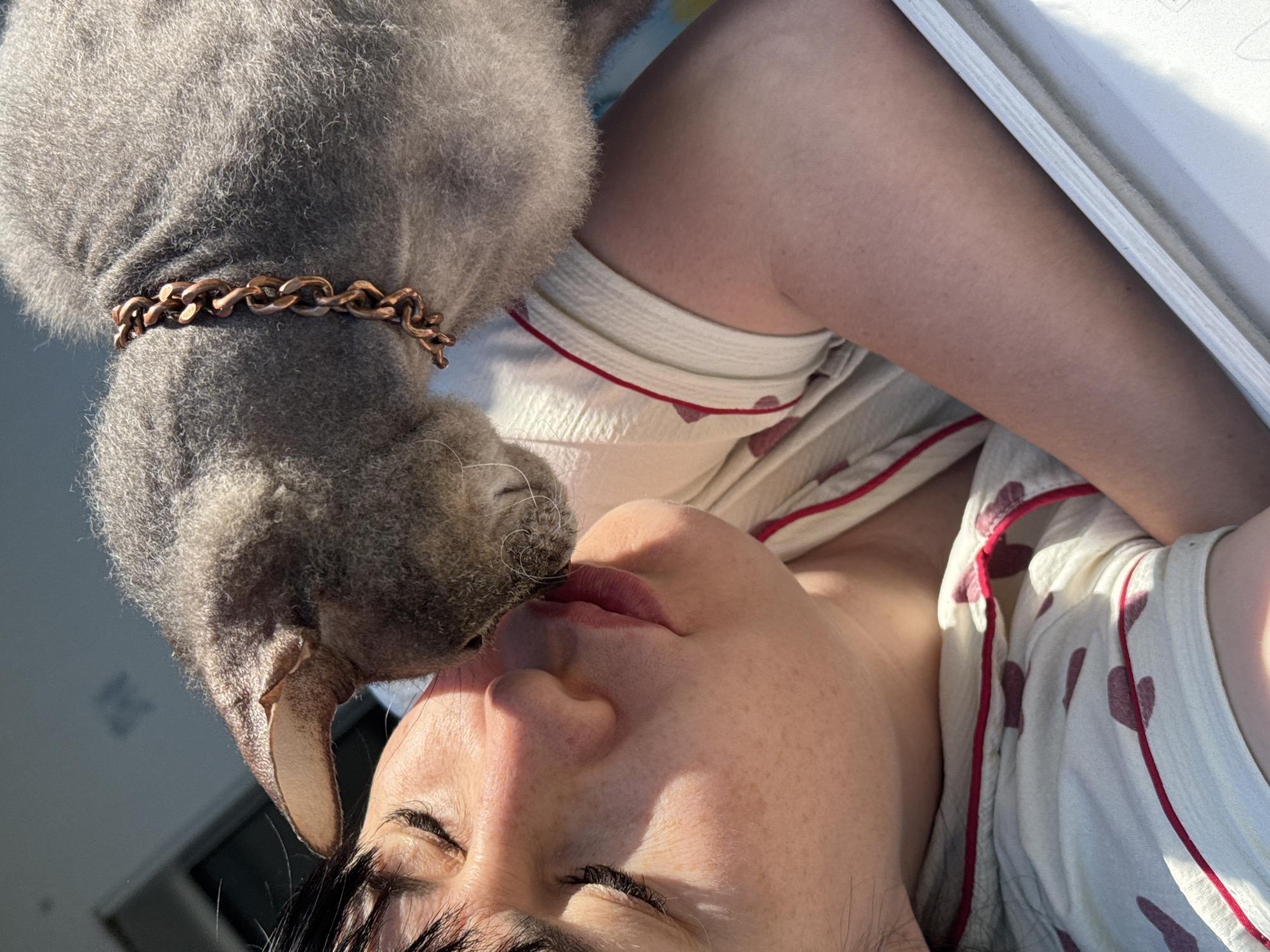 A woman is lying on her back, face tilted upward, with her eyes closed and a gray cat nuzzling her face. The cat has a copper-colored chain around its neck. The woman is wearing white pajamas with red hearts and trim. The background includes part of a white window frame and a suggestion of an exterior wall or building. It's daytime, with strong lighting. The location, Zav'yalovo, Russia, is not visually determinable from the image.