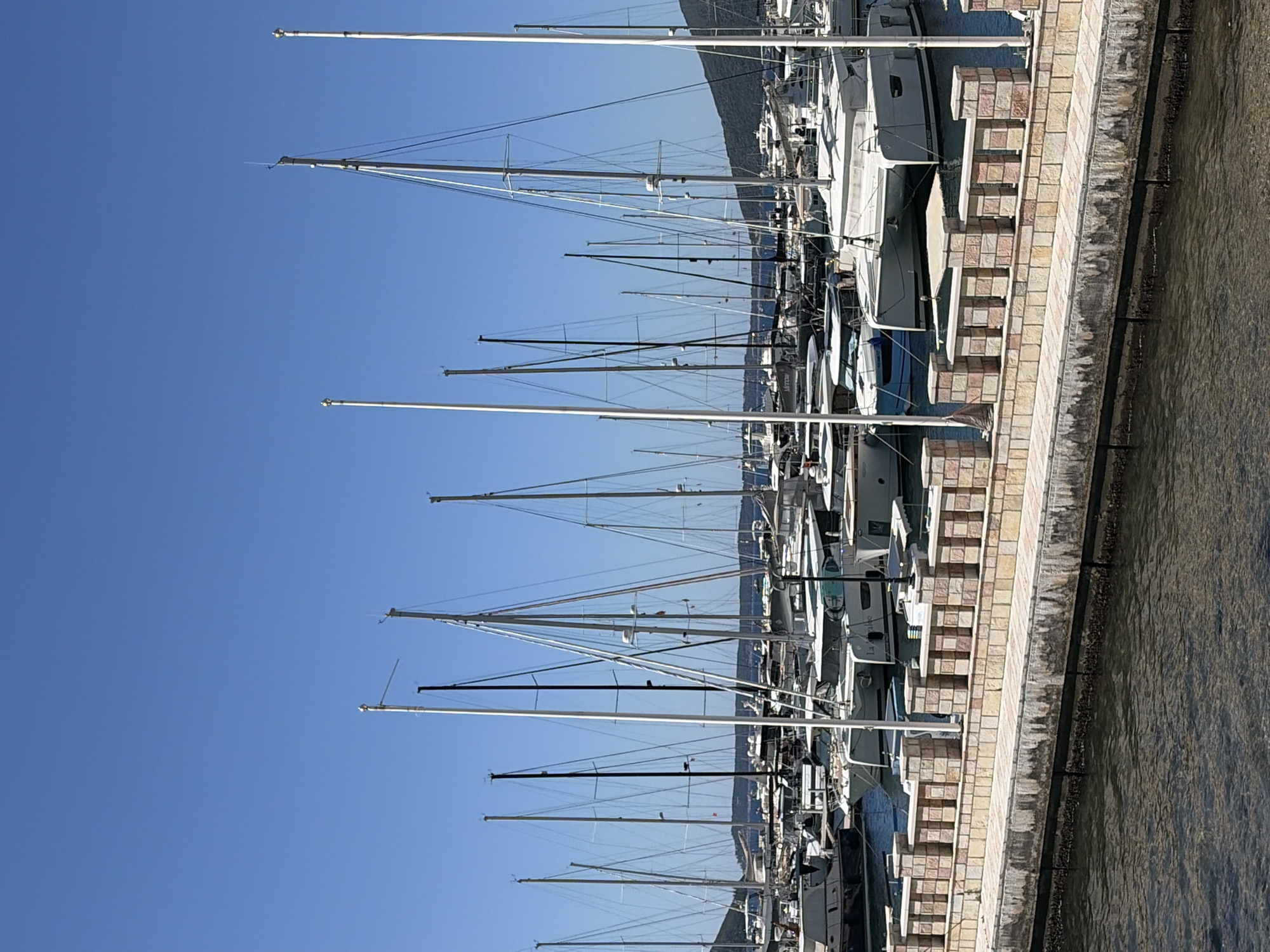 The image depicts a marina in Meljine, Montenegro, with numerous sailboats docked along a quay. The boats are aligned in a row, their masts reaching towards a clear, blue sky. The quay is constructed of stone blocks with recessed areas for docking. The water surface is visible, reflecting sunlight. There are no people visible in the frame.