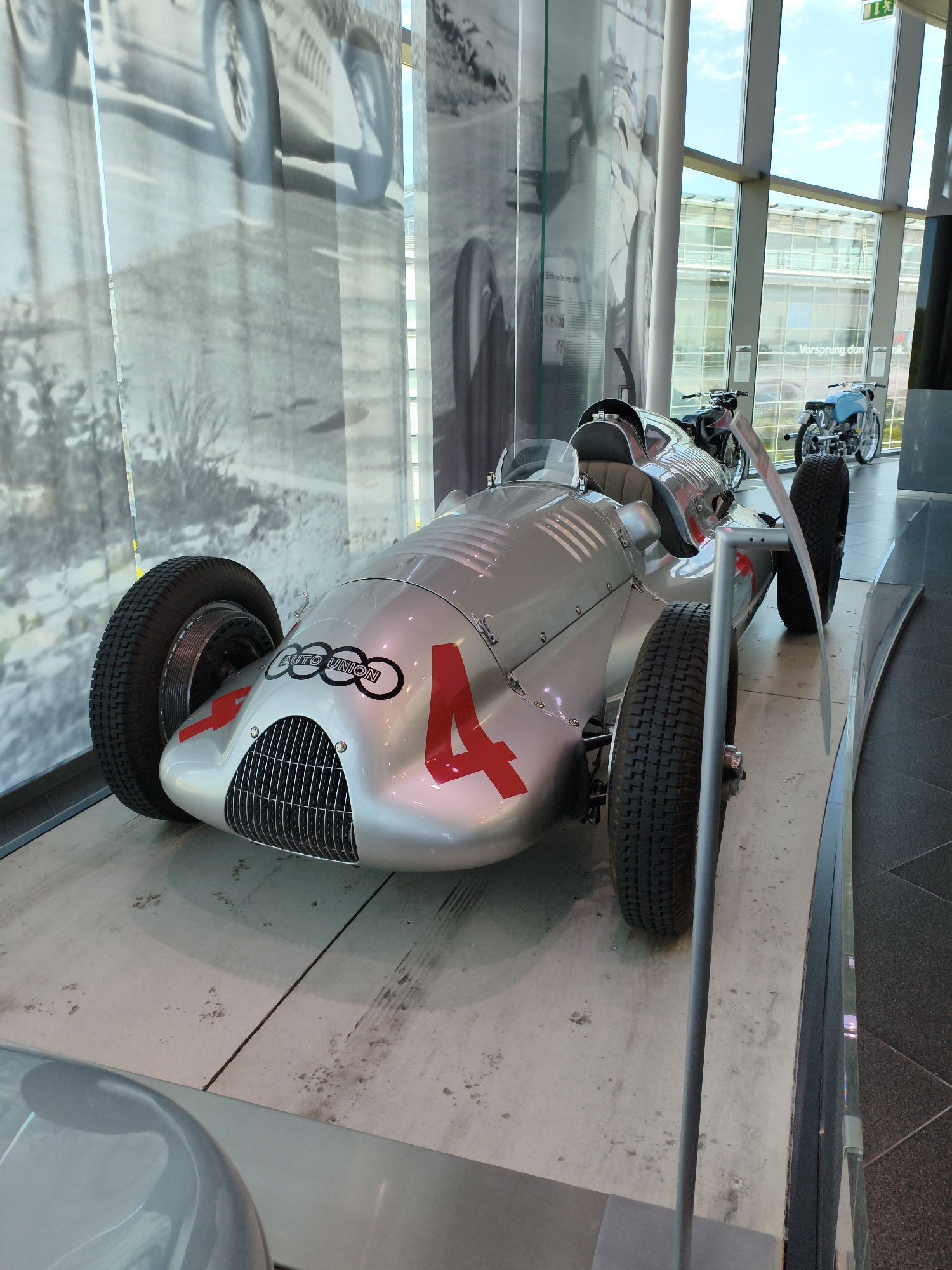 An Audi Type C racing car, silver with red accents and the number "4" prominently displayed, is exhibited indoors on a concrete floor. The car features large, treaded tires and a distinctive grille. In the background, large windows reveal a modern glass building and a blue sky with clouds. To the right, two motorcycles, one black and one blue, are also on display. The scene is a museum exhibit, likely the Audi Forum in Ingolstadt, Germany, showcasing automotive history. There are no people visible in the image.
