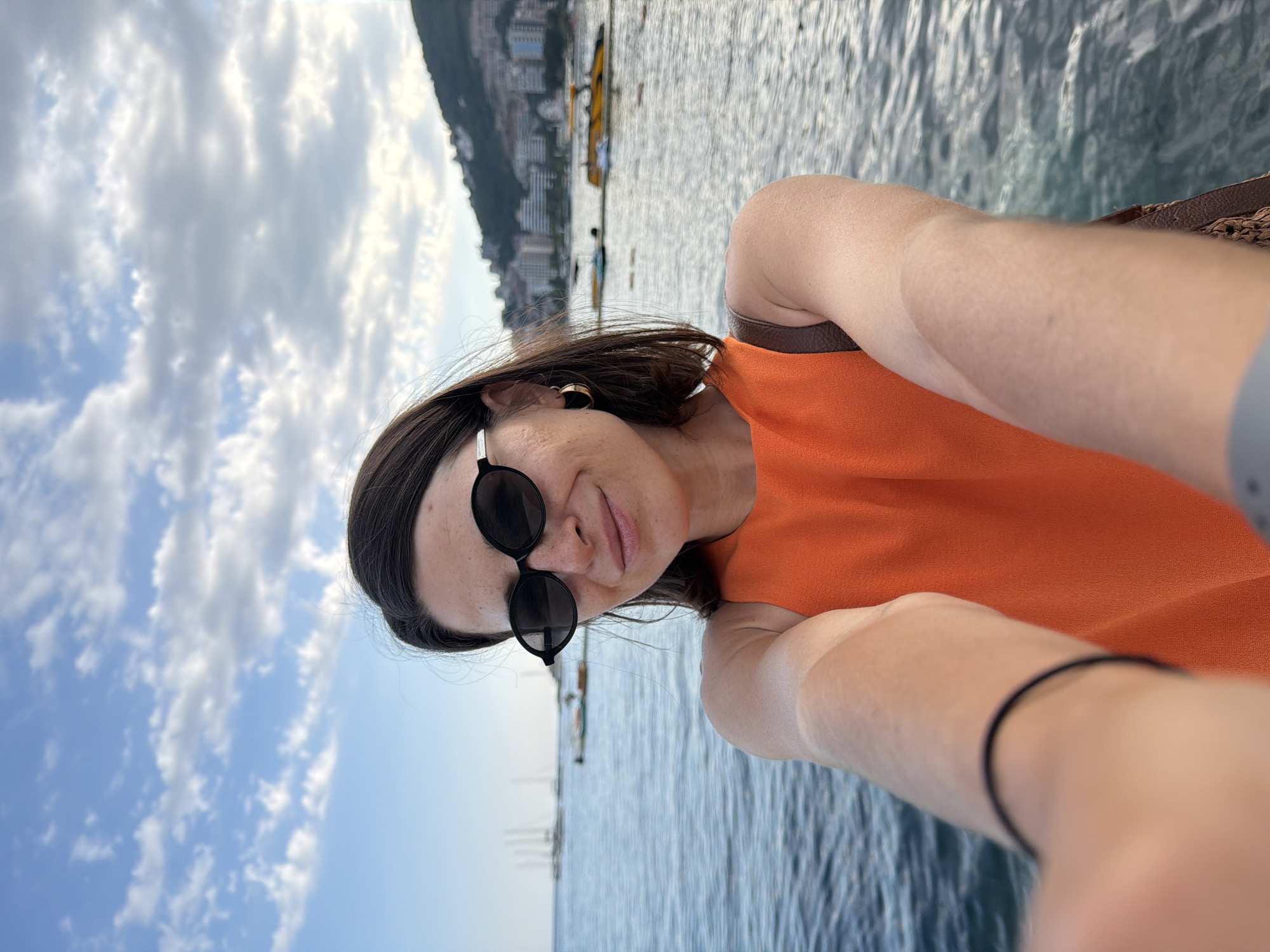 A woman is taking a selfie at the seaside in Budva, Montenegro. She is wearing an orange top and black sunglasses. The sea is calm and there are boats in the background. The sky is blue with some clouds. The city of Budva is visible in the distance on the hillside.