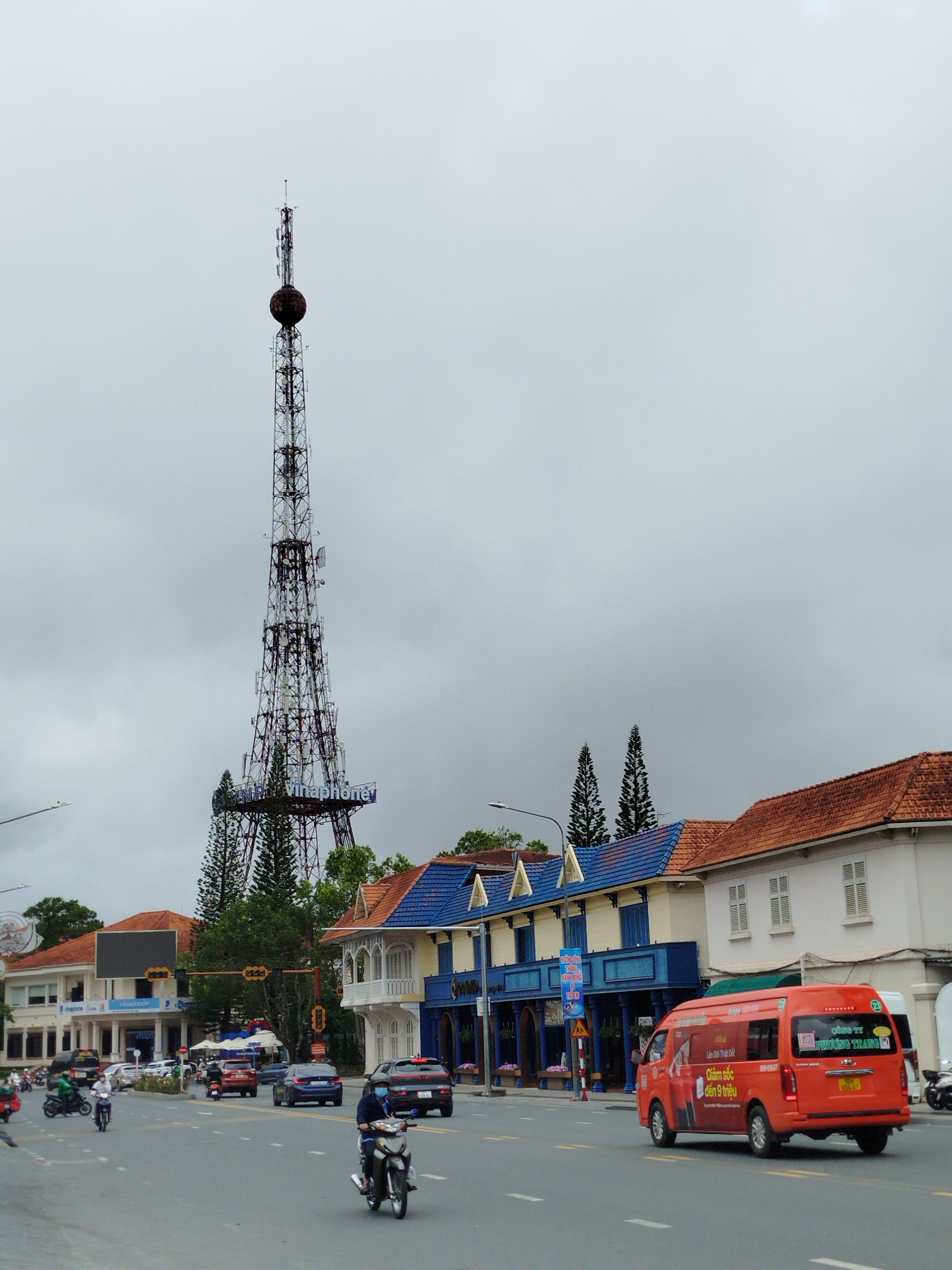 This image depicts a bustling street scene in Da Lat, Vietnam, under an overcast sky. The central feature is a tall, intricate lattice communication tower adorned with numerous antennas and a spherical structure near its peak. The word "Vinaphone" is prominently displayed in blue letters on a lower section of the tower. Two tall pine trees flank the tower's base.
The street is lined with buildings featuring distinct architecture, including red-tiled roofs and facades in white and blue. One building on the right has a striking blue roof with triangular accents and an ornate white balcony. Vehicles populate the road, including cars and several motorbikes. People are seen riding motorbikes, some wearing helmets and face masks, indicating active street life. An orange minivan, possibly a public transport or tour vehicle, moves along the right side of the road.
The time of day appears to be daytime, despite the cloudy weather. Visible text includes "Vinaphone" on the tower and on a building. The orange minivan carries text such as "Làm Đời Thật Dễ," "Giảm sốc 6 đến 9 triệu," and "CÔNG TY PHƯƠNG TRANG." Traffic lights and a large blank billboard are also visible along the street.