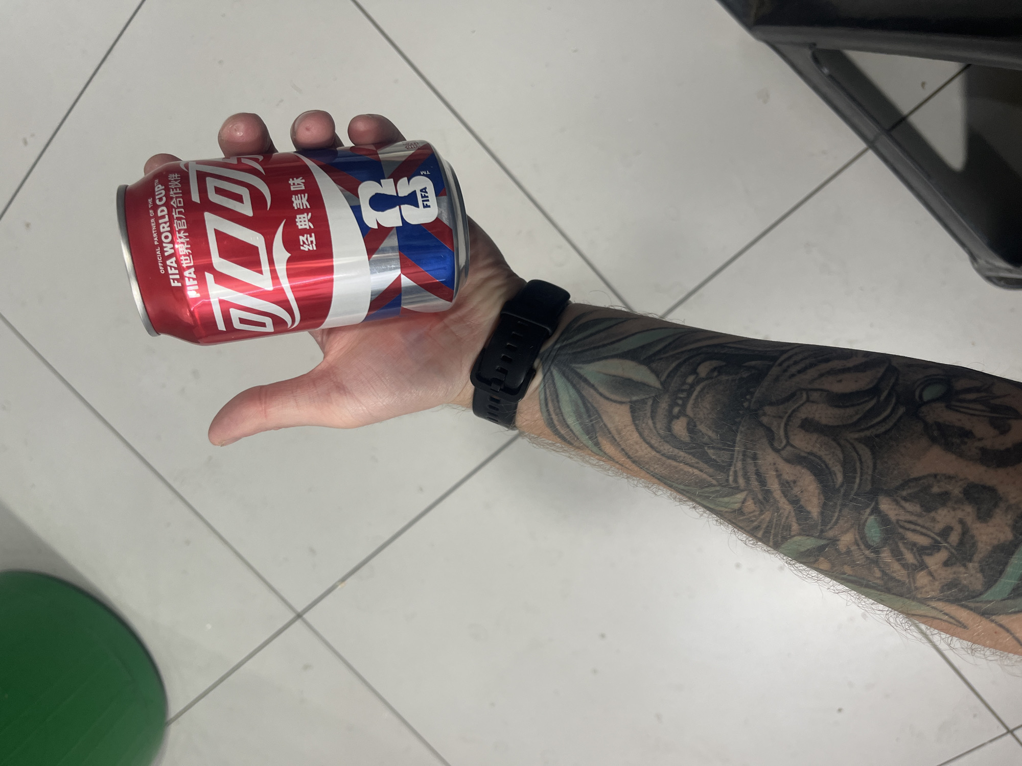 A person is holding a red and silver Coca-Cola can, which features Chinese text and FIFA World Cup branding. The can has a pattern of red and blue stripes and a FIFA logo. The person's arm, which has a tattoo of a tiger, is visible, along with a black smartwatch. The background consists of a tiled floor. The scene is indoors, possibly in a residential or commercial space. There is no indication that this image is located in Yablonovskiy, Russia.