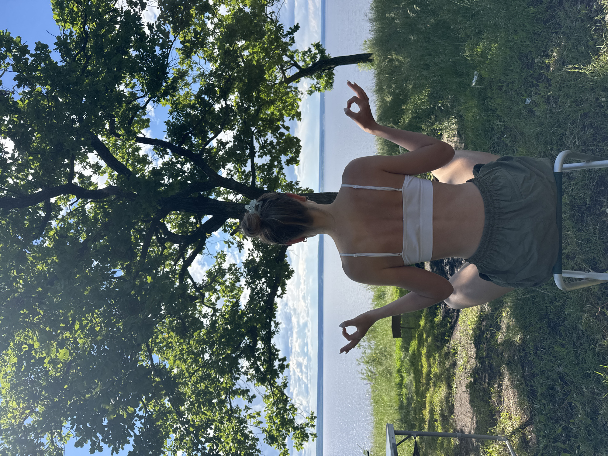 A medium shot, taken in an outdoor setting within the city of Unknown, Unknown, depicts a person from the back, seated, and engaged in a meditative practice with their hands forming "OK" gestures. A large tree with dense green foliage fills the left frame, providing a natural backdrop. Behind the person, the scene opens to a body of water, with the sky visible above it, suggesting a waterfront or lakeside location. The grassy area to the right hints at park or natural reserve. A glimpse of metallic table and legs at the bottom adds an element of human presence within the natural setting.