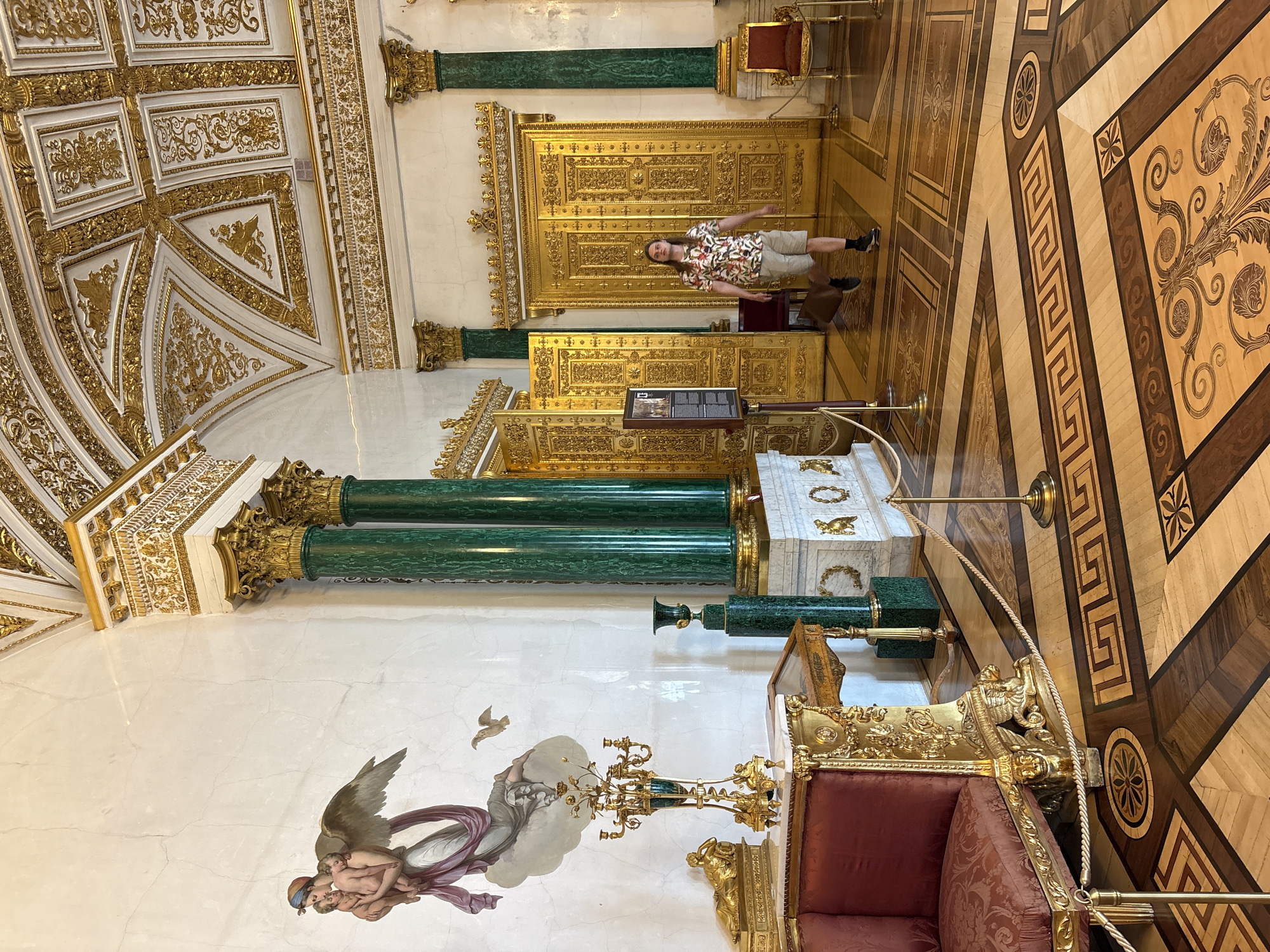 The image depicts a richly decorated interior space, likely a room within a historical building in Saint Petersburg, Russia. A man stands near an ornate, gilded door, likely part of a suite of doors or cabinets. He is wearing a short-sleeved patterned shirt and khaki shorts.
The room is characterized by opulent details: floors with intricate inlaid wood patterns, walls adorned with frescoes or murals (depicting figures with wings and clouds), and decorative architectural features such as gilded moldings and green malachite columns.
A large, gilded throne or chair is visible with a red upholstered cushion. There's also a lectern or display stand nearby with what appears to be an informational placard. Overall, the setting suggests a formal, historical, and potentially regal environment.