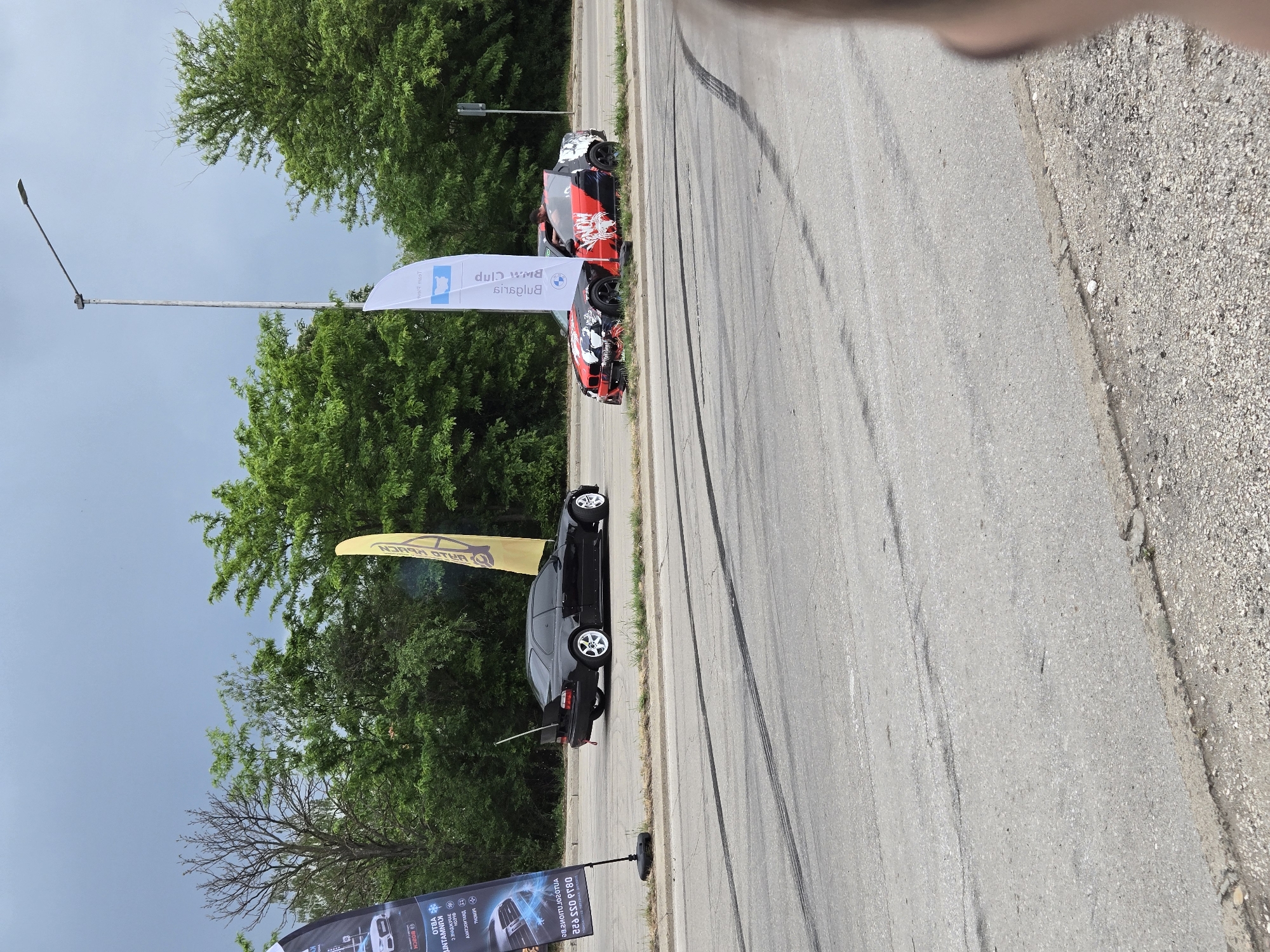 The image shows a roadside scene in an unknown location. A gray asphalt road, heavily marked with tire skid marks, dominates the right side of the image. The road's edge transitions into a gravelly shoulder. A line of trees forms a backdrop on the left side. Several cars are parked near the trees: a black sedan and a red and black car. Banners stand near the cars, but the words are blurry. A streetlight is visible near the trees. The sky is overcast.