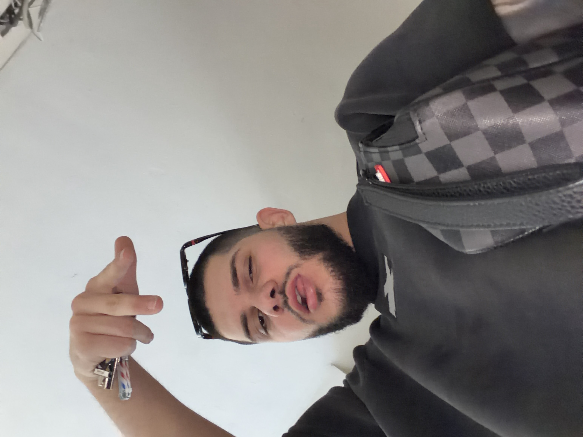 A man is depicted in a selfie. He is holding keys in his left hand and gesturing with his fingers. He has a beard, and eyeglasses are perched on his head. He is wearing a dark grey hooded sweatshirt and a gray checkered bag is slung across his chest. The background is a plain white wall. The location is unknown and there is no evidence of this image being in Sofia, Bulgaria.