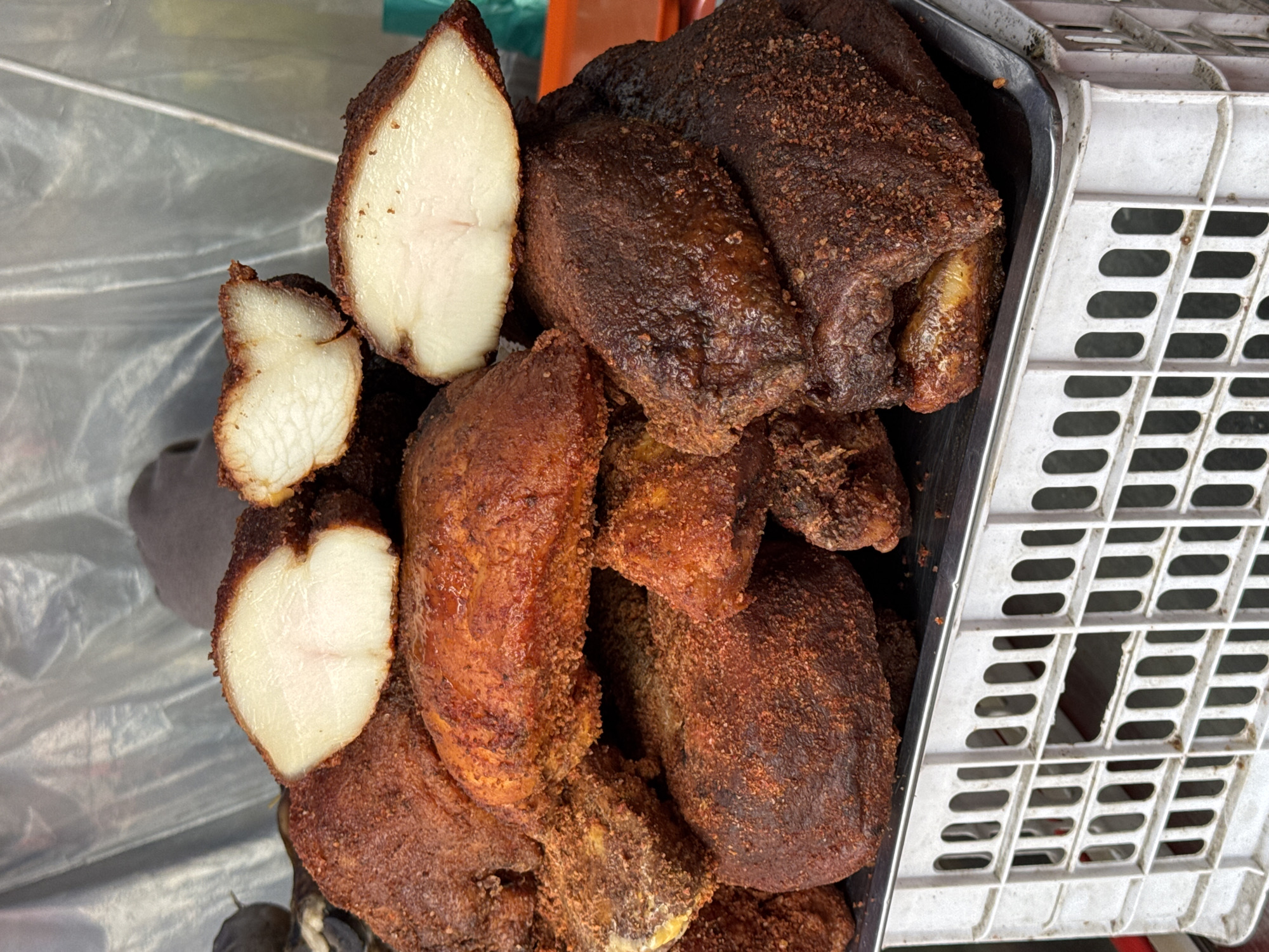 The image presents a close-up view of numerous pieces of deeply roasted or fried food, piled within a rectangular metal tray. The food items feature a dark, reddish-brown, heavily seasoned exterior, appearing somewhat crispy or crusted. Several pieces are cut in half, revealing a pale, off-white interior with a fibrous texture, strongly suggesting cooked poultry, possibly chicken or duck breast. Some subtle pinkish hues are discernible within the pale interior.
The metal tray containing the food is positioned next to a white plastic utility crate, which is partially visible on the right side of the frame, showcasing its distinctive grid pattern. The background includes a translucent plastic sheet or bag, which, along with the utility crate and the presentation of the food in bulk, indicates a commercial setting such as a market stall or food vendor.
The scene likely depicts food prepared for sale in Bishkek City, Kyrgyzstan. There are no people visible, and no discernible text. The lighting is bright, suggesting daytime, but specific weather conditions or the exact time of day cannot be determined. The image focuses entirely on the food and its immediate containers, conveying a sense of hearty, prepared meal options.