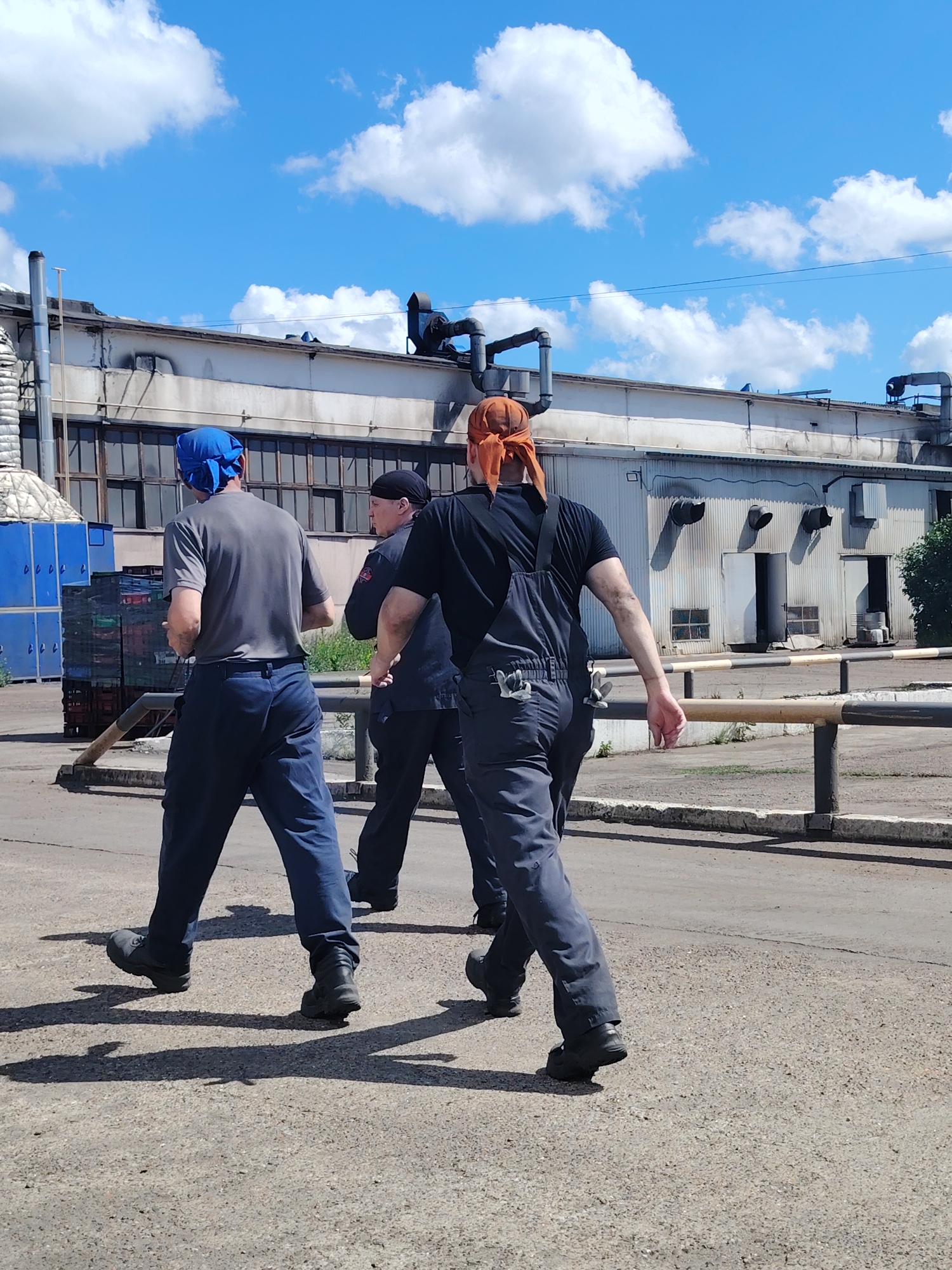 Three men in work clothes are walking away from the camera on a paved surface in front of a large, industrial building. The man in the foreground on the right is wearing dark overalls and an orange bandana. The man in the middle is wearing a dark uniform and a black cap. The man on the left is wearing a gray t-shirt and blue pants with a blue bandana. The building appears to be a factory or warehouse, with large doors and windows. There are stacked blue crates to the left of the building. The sky is blue with white clouds. The location is identified as Noviy, Russia.