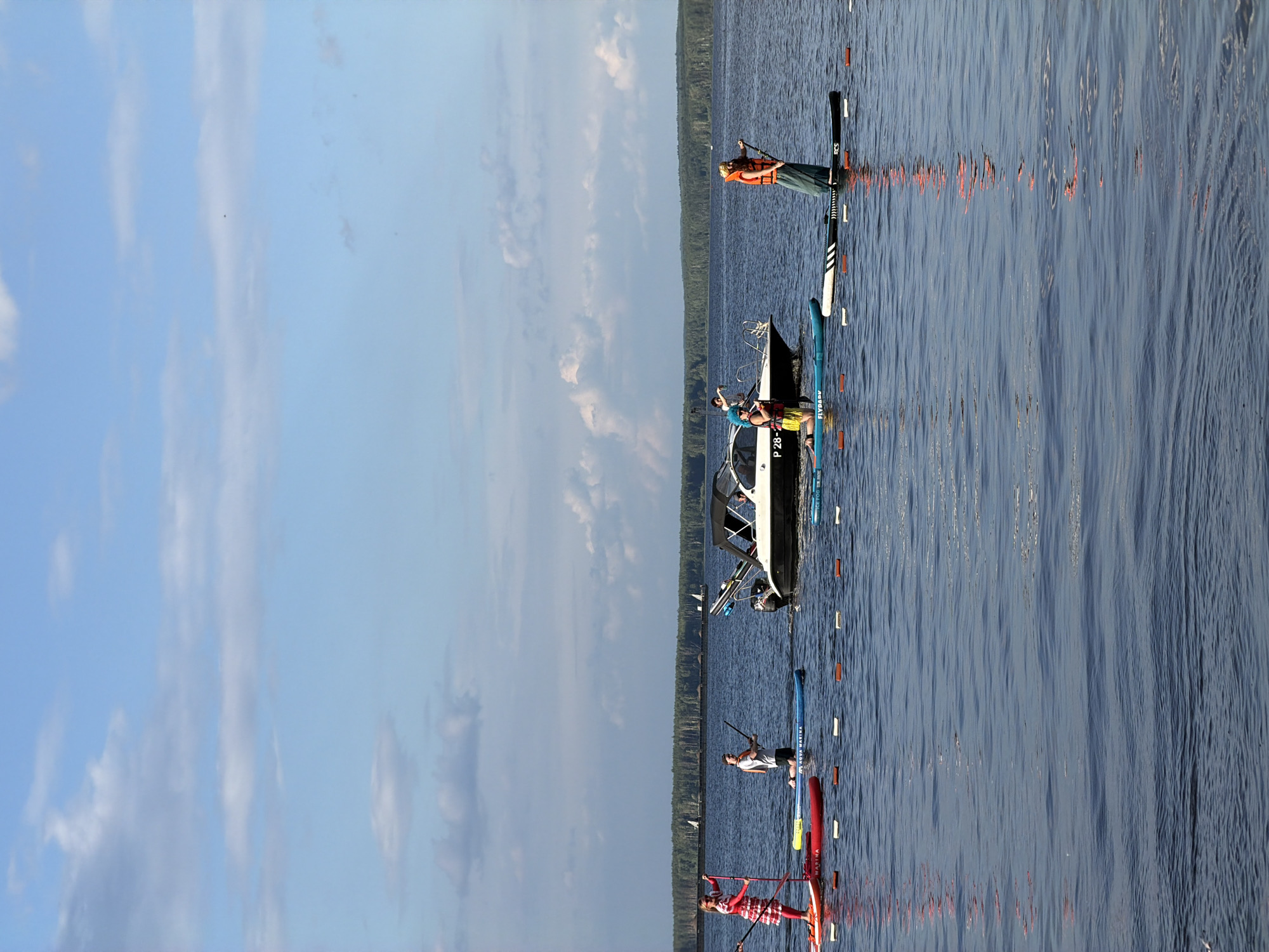 This is a photograph taken on a sunny day at the Kama River in Chaikovsky, Russia. The scene depicts several people engaged in stand-up paddleboarding on the calm, blue water. In the foreground, two people are on paddleboards, one in a red and white dress and the other in a white t-shirt and grey shorts, holding paddles. To the right, a motorboat is docked or slowly moving, with at least two individuals visible on it. Further to the right, another person is standing on a paddleboard wearing an orange life vest and a long green skirt. In the background, a dense line of trees forms the horizon, with a few distant sailboats also visible. The sky is a clear blue with scattered white clouds. The water is reflecting the sky and the surrounding environment. The image captures a recreational activity on a wide river.