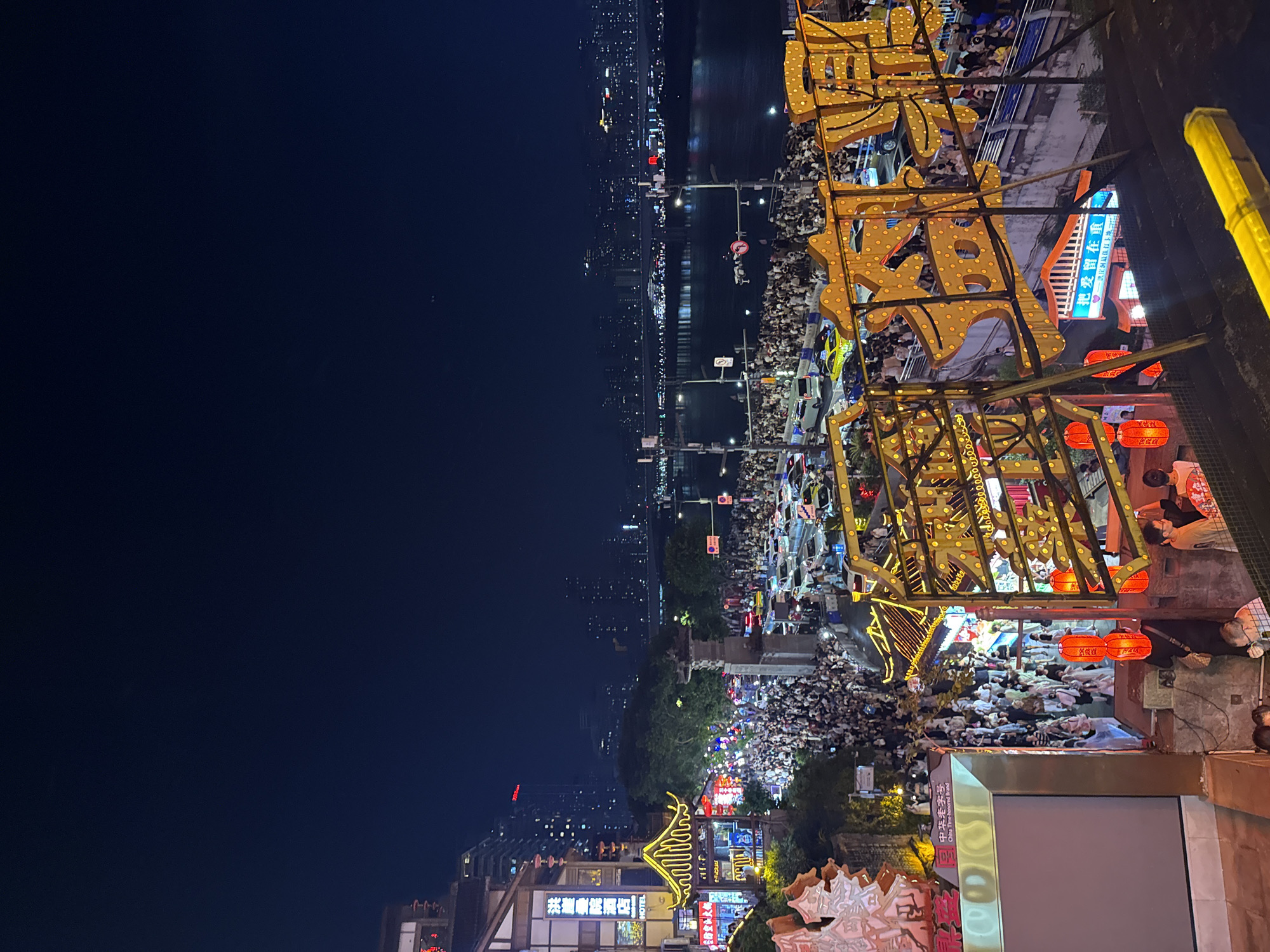 This image captures a vibrant nighttime scene in Yuzhong District, Chongqing, China, showcasing a lively pedestrian street lined with shops and illuminated by a profusion of lights. The architecture features traditional Chinese elements, with ornate, yellow-lit eaves and red lanterns adorning the buildings. A dense crowd of people fills the street, appearing to be engaged in leisurely activities, with many looking towards the shops and the opposite bank of the river, which is illuminated by the city lights. The street is also bustling with vehicular traffic, including cars and taxis, suggesting a popular and active urban area. In the background, the city skyline rises impressively, with numerous buildings and lights reflecting on the calm river. The overall atmosphere is one of festive energy and urban dynamism.
