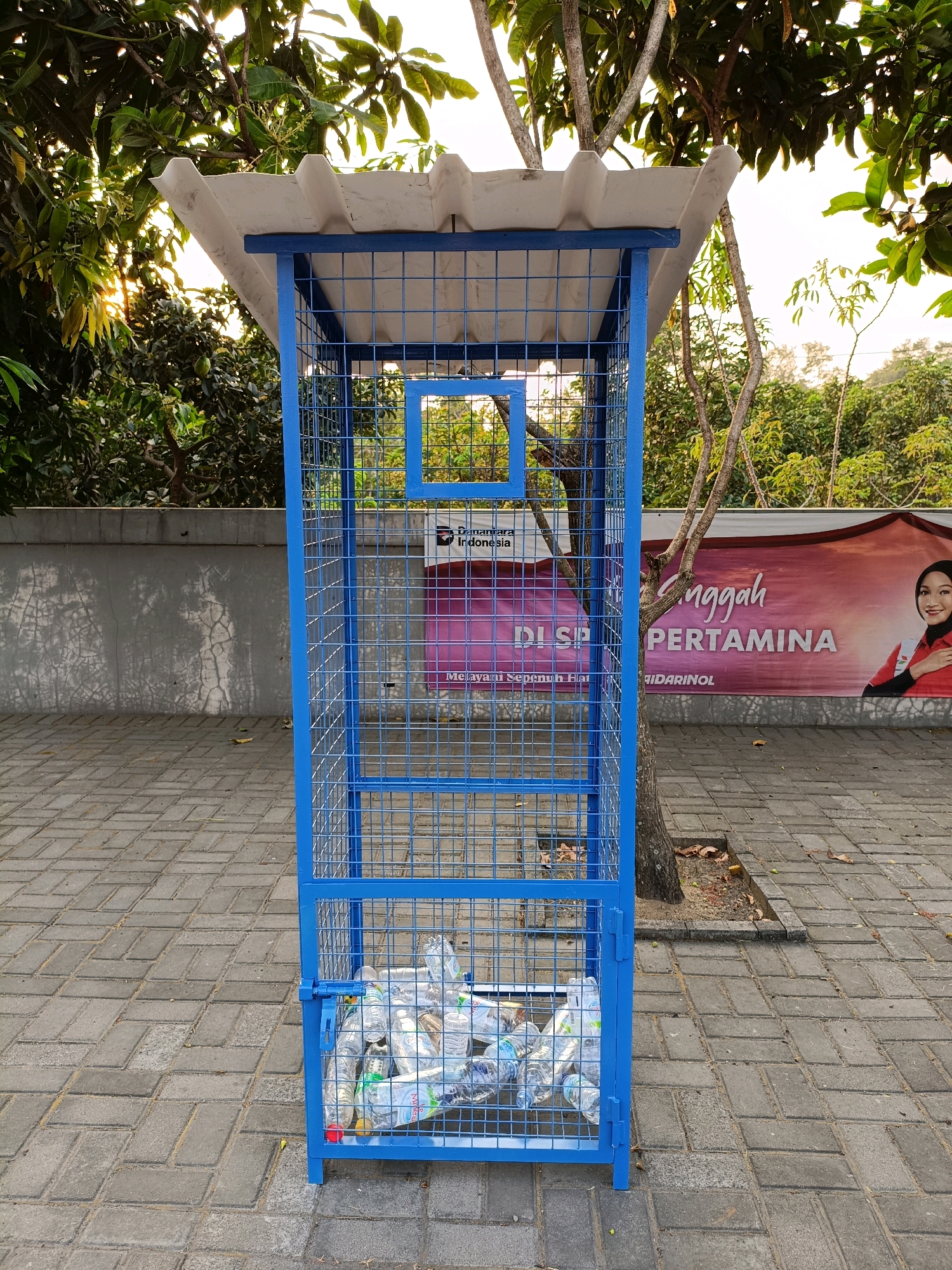 This is a medium-shot, eye-level, outdoor photograph of a blue metal cage recycling bin filled with plastic bottles. The bin is positioned on a paved area with a patterned brick surface. Behind the bin, there is a banner featuring text and an image of a woman. Trees with green leaves are visible in the background, and sunlight filters through the branches. The scene appears to be in Maguwoharjo, Indonesia. The bin has a white, corrugated roof and a small square opening on its front. The recycling bin has a metal latch to secure its door.