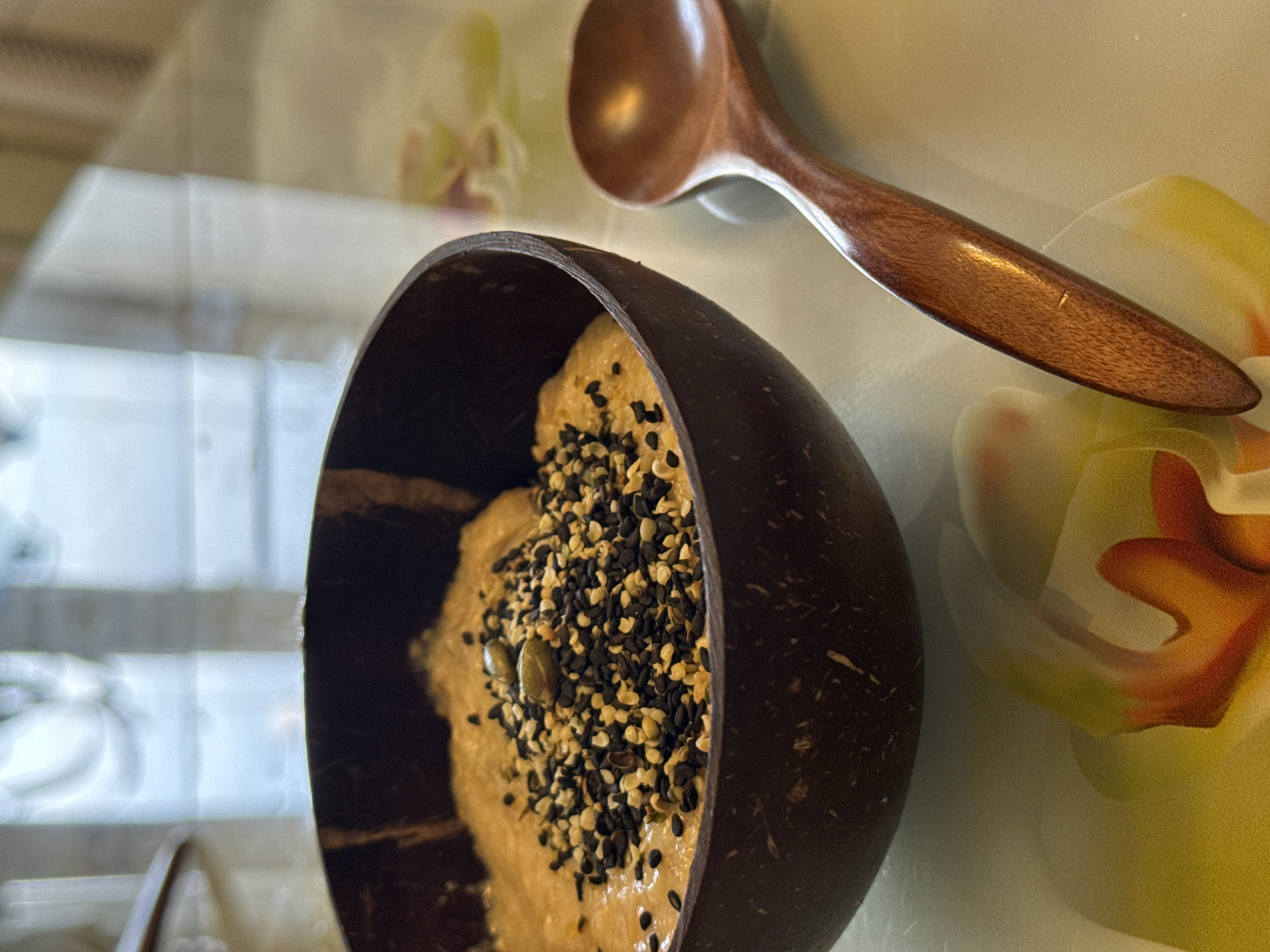 A close-up shot depicts a wooden bowl filled with a light-colored substance topped with a mixture of black and white seeds, and a few green pumpkin seeds. A wooden spoon rests to the side of the bowl. The scene takes place on a surface with a floral pattern. The location is in the city of Unknown, Unknown.