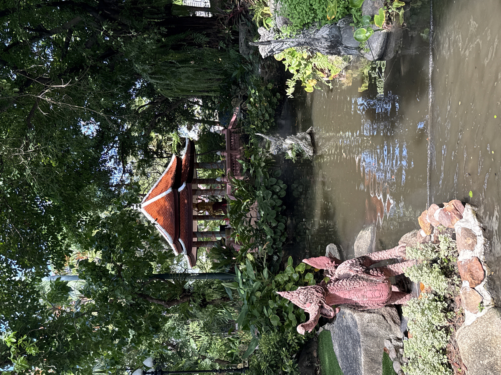 The image shows a pond in a garden setting, located in the Samrong Nuea Subdistrict Municipality, Thailand. The pond's water is murky. At the edge of the pond is a pink dragon statue on a rock formation with a small green grass border. On the opposite side of the pond, there is a gazebo with a red-tiled roof. The scene is surrounded by dense green foliage, with trees and plants obscuring parts of the pond and gazebo. The lighting indicates daylight conditions.