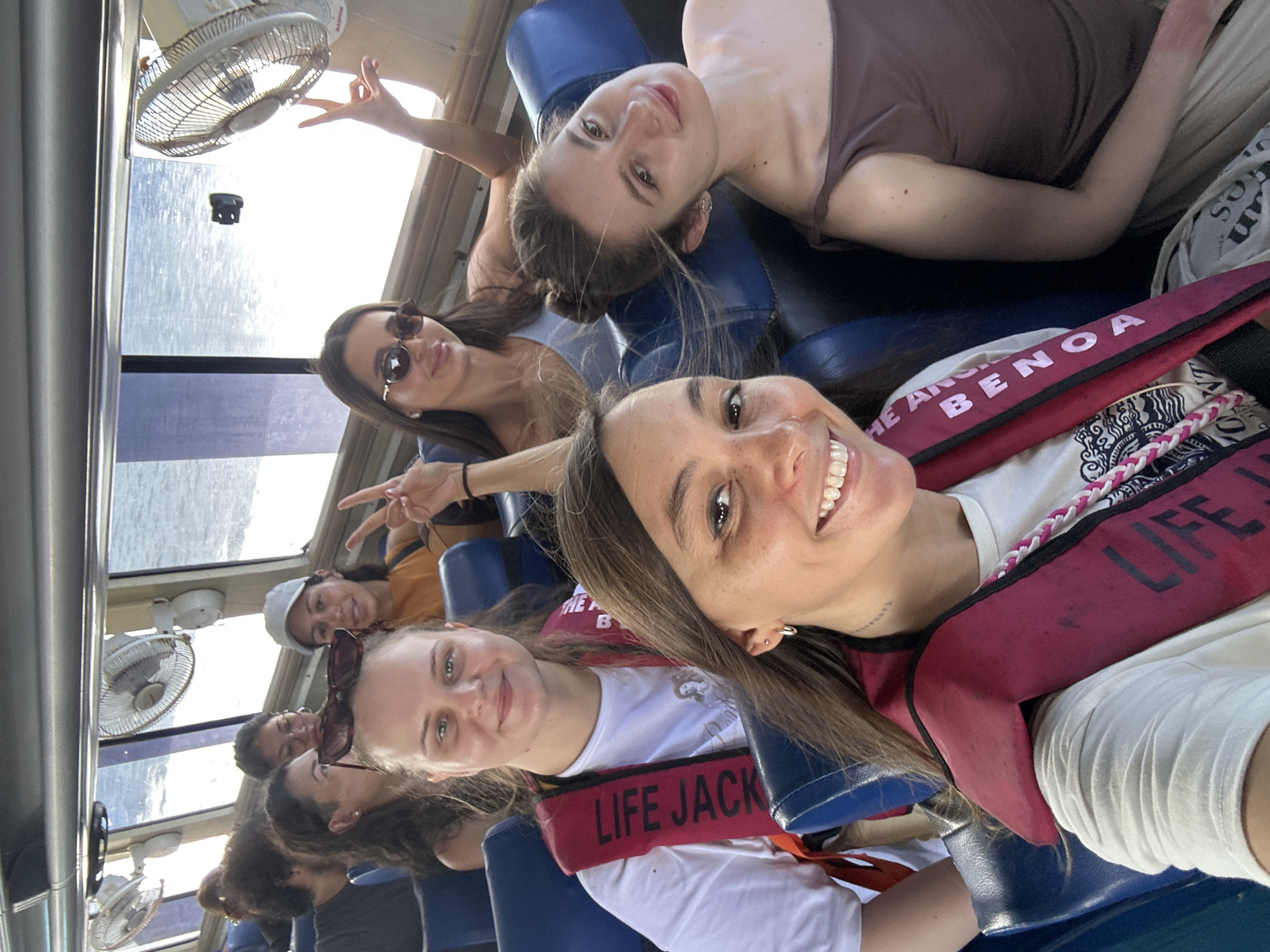 A group of approximately seven women are inside a boat or enclosed vehicle, likely on a tour or excursion. They are wearing life jackets, and some are posing for a photograph. The location is unspecified, but the presence of a boat and life jackets suggests proximity to water. The photograph is taken from an unusual angle, potentially by one of the women in the group. The overall environment appears bright and sunny.
