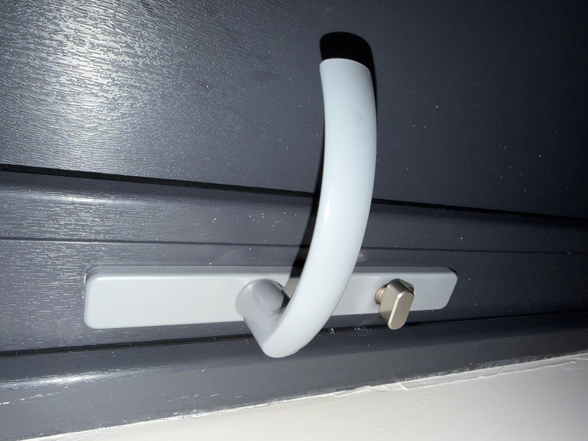 A close-up, eye-level, vertical shot shows a grey door handle and lock mechanism on a dark grey door. The door has a vertical wood grain texture. The door handle is a light grey, curved bar with a rounded end that extends horizontally. A silver, oval-shaped lock is located below the handle. The image is cropped, showing only the door and its hardware. The scene appears to be an interior door, possibly in an apartment building. The white wall to the left of the door is slightly out of focus. This image was taken in Szczecin, Poland.