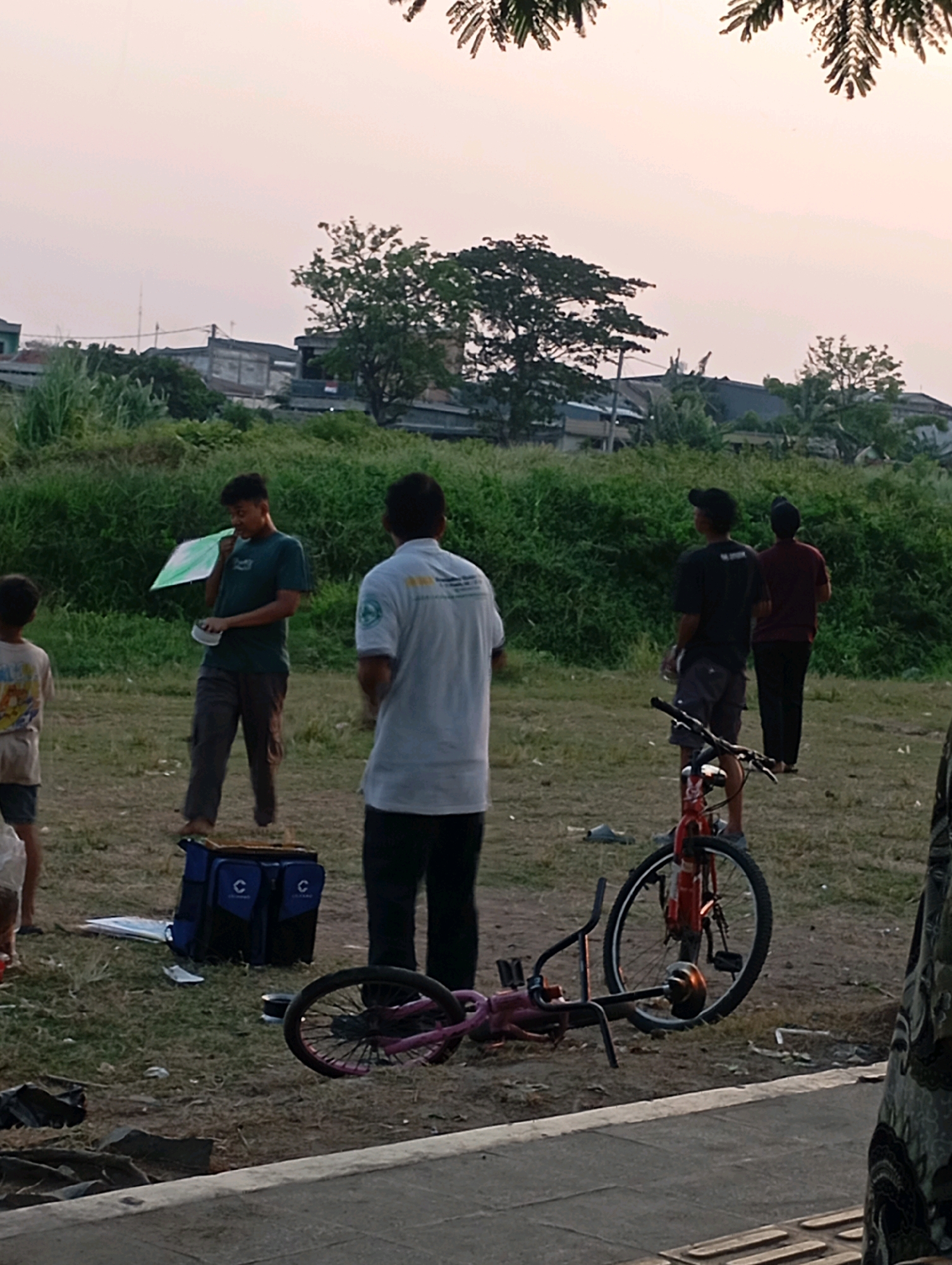 In Babelan, Indonesia, a group of people are gathered in a grassy, open area. A young man in a dark green t-shirt and dark pants holds a green and white kite, appearing to be preparing it. Beside him, a blue and black bag with "C" logos sits on the ground. To his left, a young child in a light-colored t-shirt stands. In the background, a man in a white t-shirt and black pants stands facing away from the camera. Further back, two other men, one in a black t-shirt and shorts, the other in a maroon t-shirt and dark pants, are also visible. A red bicycle is lying on its side in the foreground, with its wheels facing upwards. The environment appears to be a park or open field with lush green vegetation and some buildings visible in the distance. The sky is a pale, hazy color, suggesting it may be dusk or dawn.