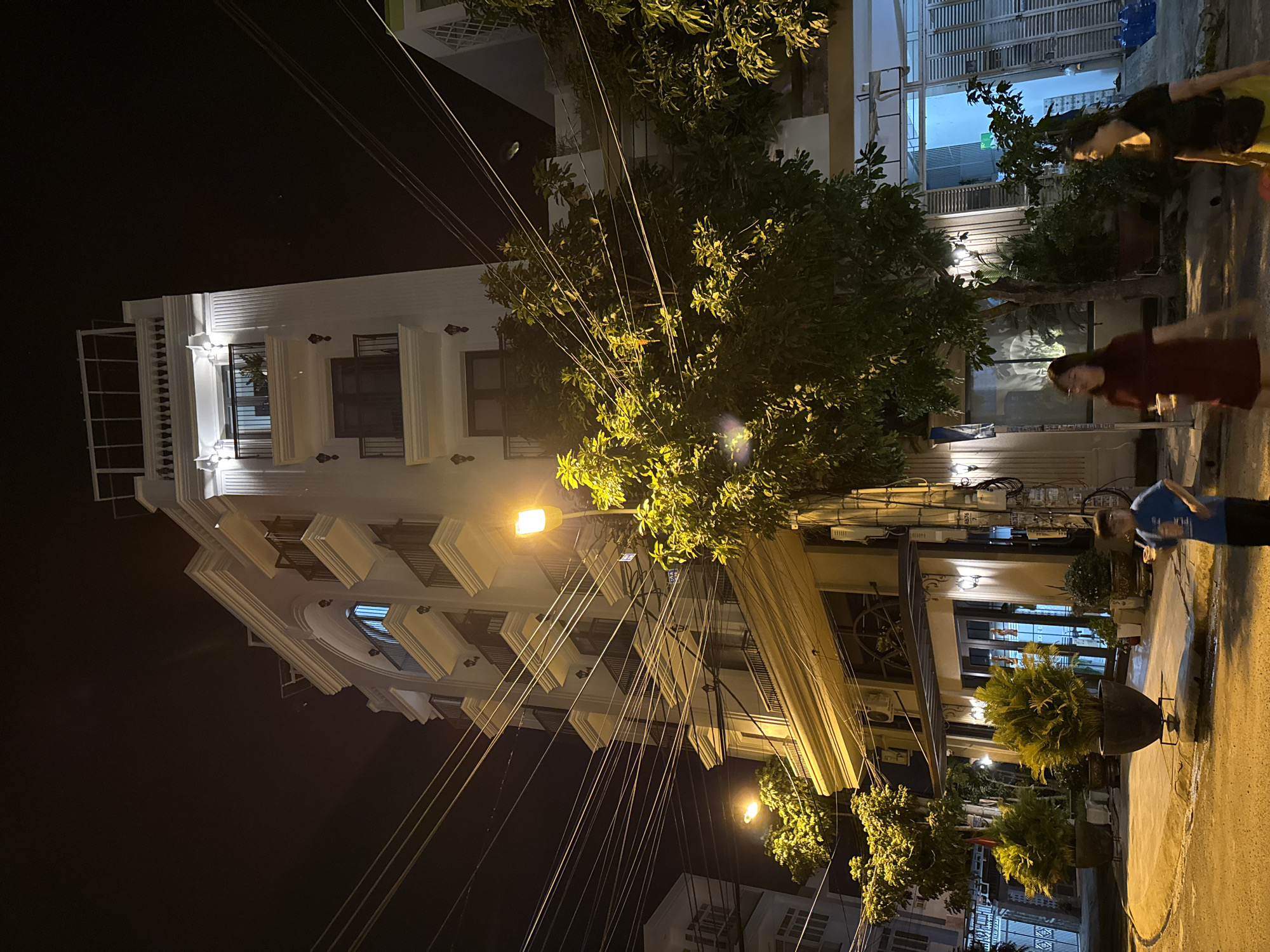 The image depicts a street scene at night in Bac Nha Trang Ward, Vietnam. A multi-story white building with balconies and illuminated windows dominates the background. Numerous tangled electrical wires are prominently strung across the foreground, obscuring parts of the building and the sky. Several trees with green foliage are visible, some illuminated by streetlights. Two women and a young boy are present. One woman, wearing a dark top and yellow skirt, is walking towards the right. Another woman, in a maroon dress, is holding a cup and walking towards the left. The boy, wearing a blue shirt and black pants, is also walking towards the left. The ground appears to be asphalt or concrete, with some street lights casting a warm glow. Potted plants and outdoor lighting are also visible around the building's entrance.