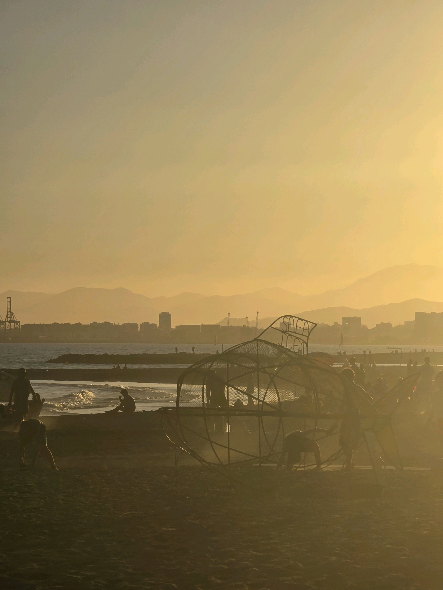 The image captures a coastal scene at sunset in Málaga, Spain, likely from the area near the port or the El Palo district, given the presence of industrial cranes and the mountainous backdrop typical of the region. The sky is a hazy, warm yellow-orange, indicating the sun is low on the horizon and casting a diffused light over the entire scene.
In the foreground, a beach is visible, with dark sand and the subtle texture of the grains evident. Several people are scattered across the sand and at the water's edge. Some are silhouetted against the bright sky, making their individual features indistinct. One individual is seated on the sand near the water, and another appears to be walking towards the left. A large, skeletal structure resembling a giant fish or marine creature, made of metal tubing and mesh, dominates the mid-ground. Several people are interacting with or near this structure, with some appearing to climb on it or move around its base.
Further back, the sea is calm, with gentle waves lapping at the shore. A breakwater or pier extends into the water, and a few figures are visible on it. Beyond the water, the city skyline is a soft, muted silhouette against the hazy sky. Prominent industrial cranes are visible to the left of the city, suggesting proximity to the port. The distant landscape is defined by layered, soft-focus mountains that fade into the atmospheric haze. The overall impression is one of a relaxed, late-afternoon or early-evening beach environment, with a large art installation or playground feature as a focal point.