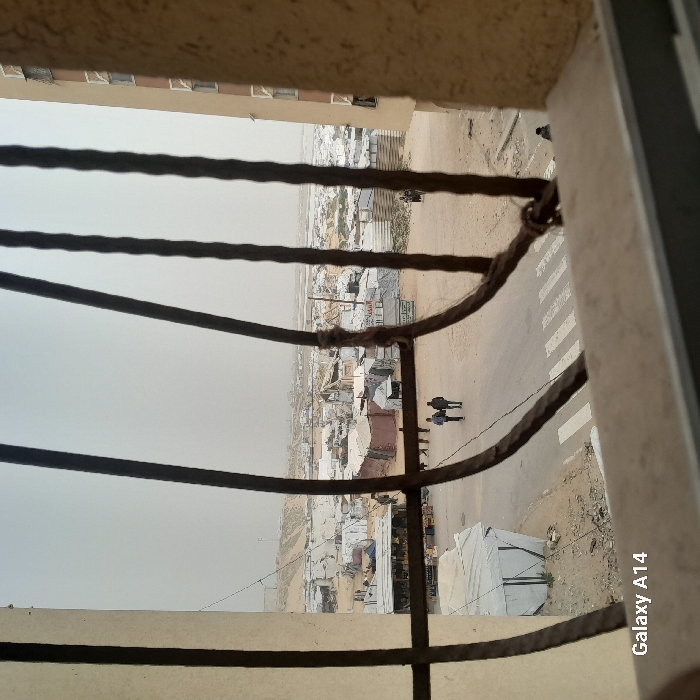 An upward-angled view through a balcony fence shows a sprawling settlement or refugee camp with makeshift buildings. The landscape is dusty, and the sky is overcast. A road with a zebra crossing cuts through the area. People walk on the roadside. A tall building is partially visible above the fence, suggesting the photograph was taken from a higher level. The "Galaxy A14" watermark is visible in the lower right corner.