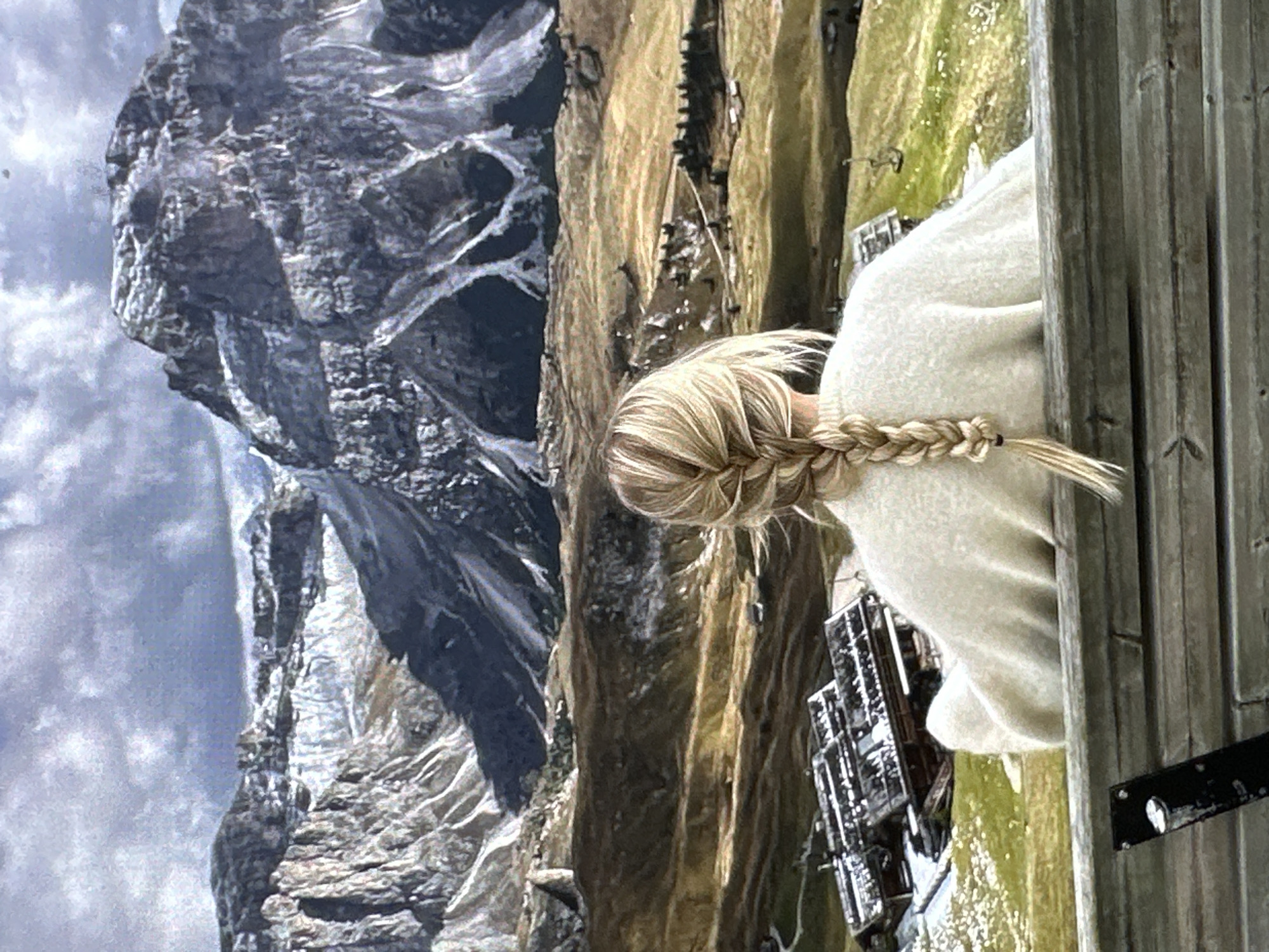 A woman with blonde hair in a braid faces a mountain scene. The mountain is craggy and partly covered in snow. Below the mountain is a sloped, grassy and rocky area with buildings. The woman stands near a wooden structure. The location is unknown.