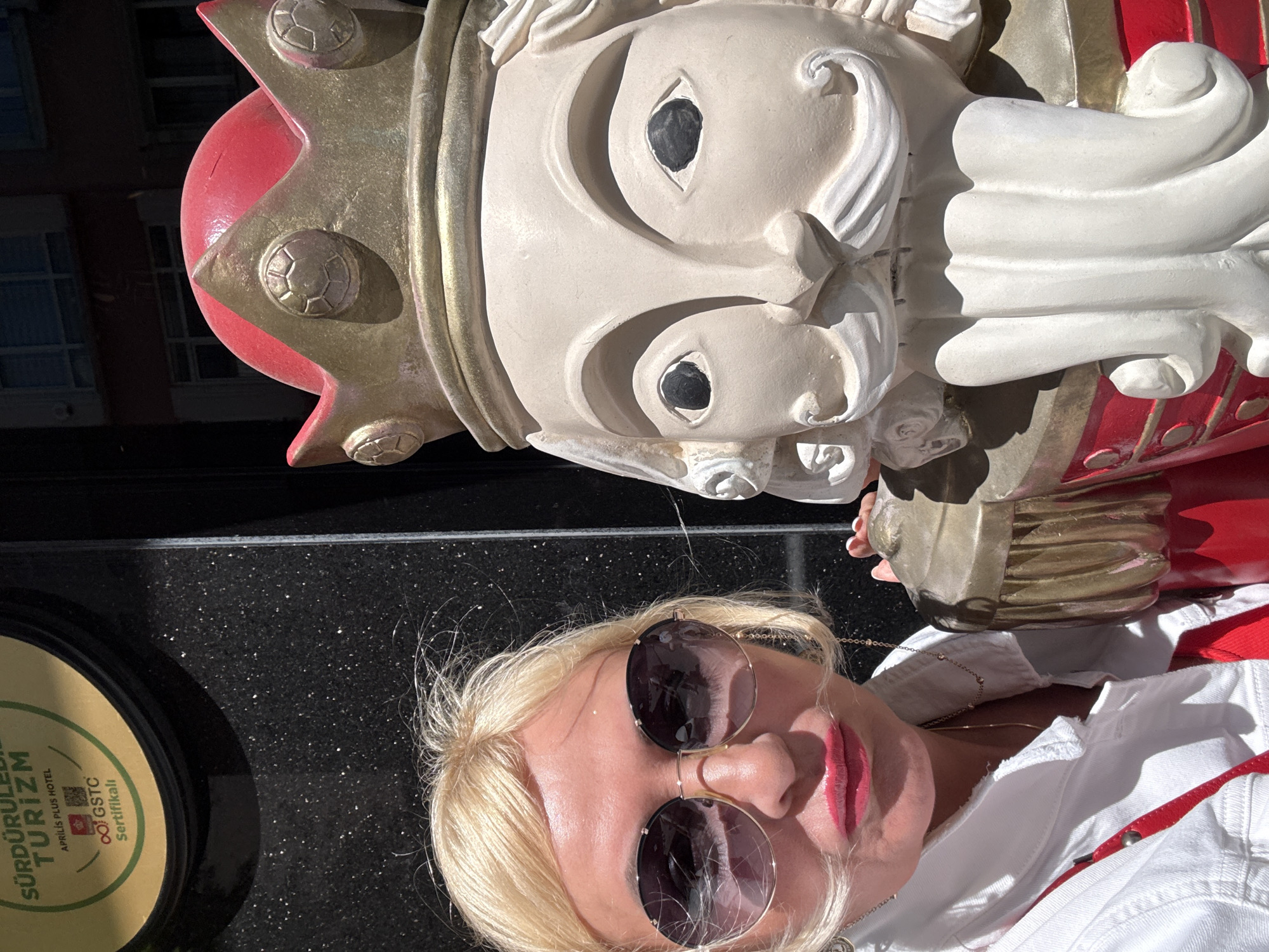 A portrait-style shot depicts a blonde woman with sunglasses posing next to a statue of a king. The woman, who is positioned at the bottom of the frame, is wearing a white jacket and multiple necklaces. The statue, dominating the upper half of the image, is a regal figure with a crown and ornate details. To the left of the woman, a circular sign with the words "SÜRDÜRÜLEBİLİR TURİZM" and "APRILIS PLUS HOTEL" is partially visible. The location is identified as being in the city of Unknown, Unknown.