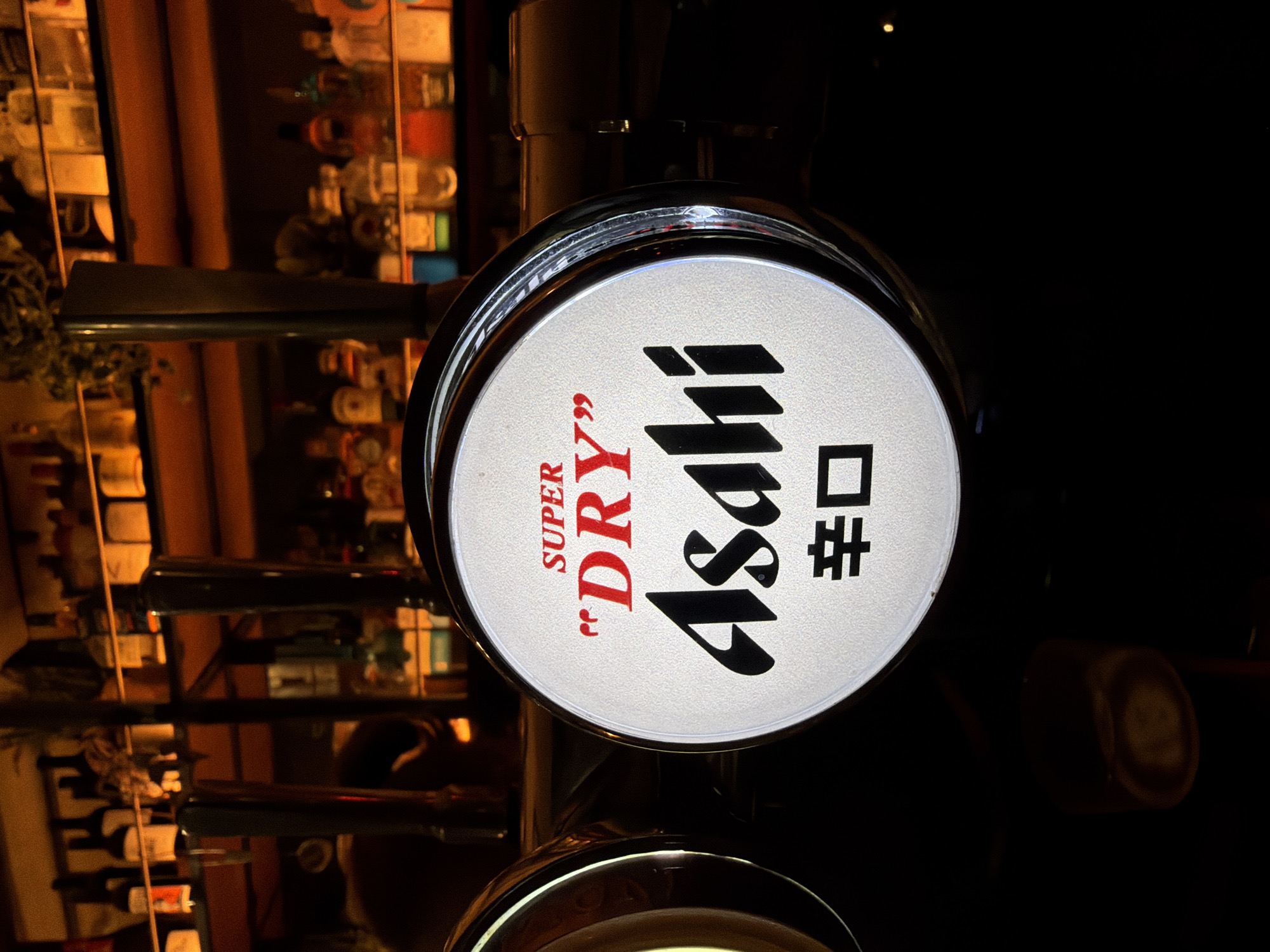 The image depicts an Asahi Super Dry beer tap in a dimly lit bar. The tap features a circular white face with the words "SUPER 'DRY'" in red and "Asahi" and Japanese characters in black. The background shows shelves stocked with various bottles, likely liquor, suggesting a bar setting. The location, city, and country cannot be determined from the image.