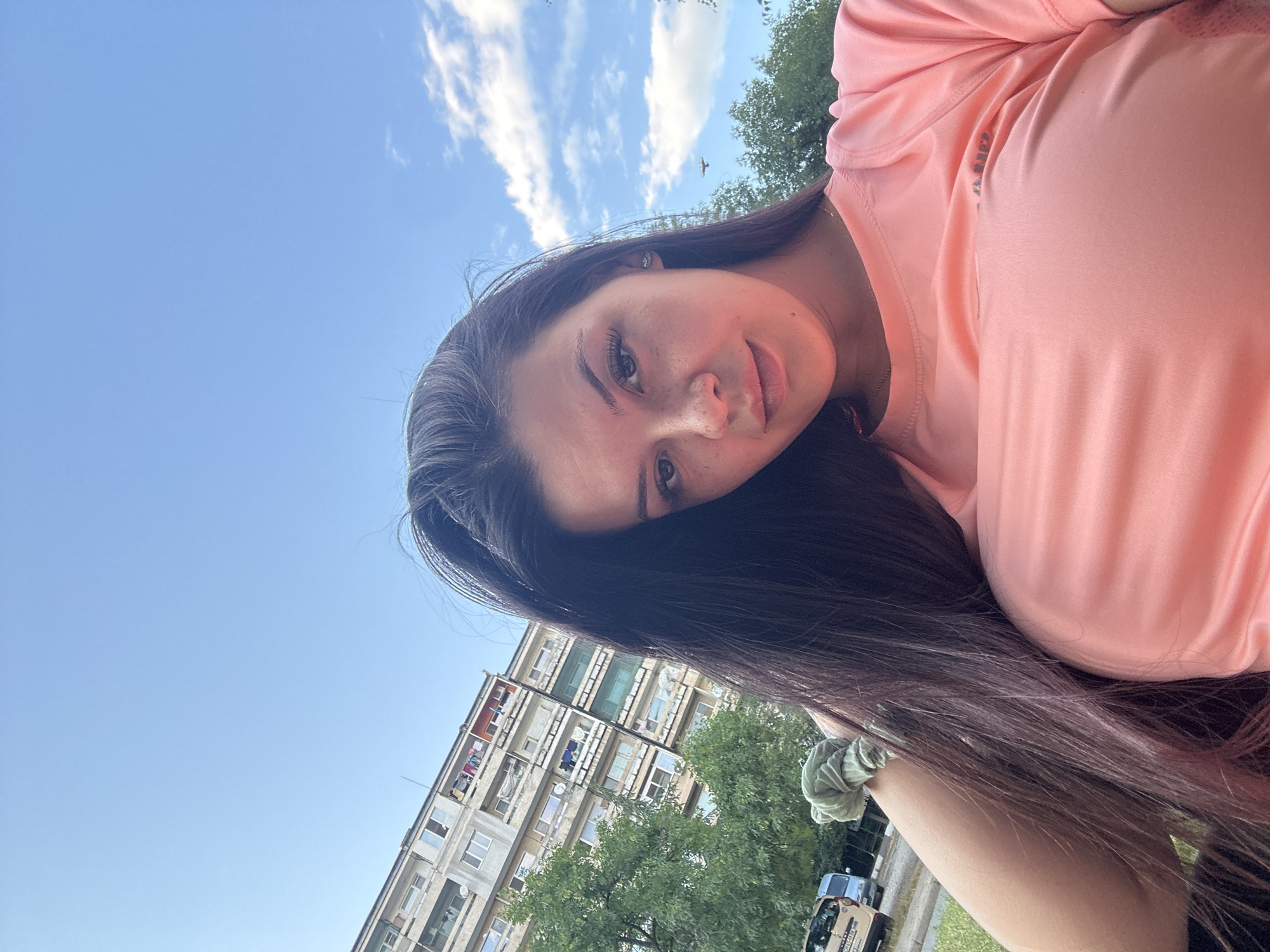 A woman is taking a selfie outdoors on a sunny day with a blue sky and some clouds. She has long dark hair and is wearing a coral-colored t-shirt. In the background, there is a multi-story apartment building, trees, and a street with parked vehicles. The location is the city of Sliven, Bulgaria.