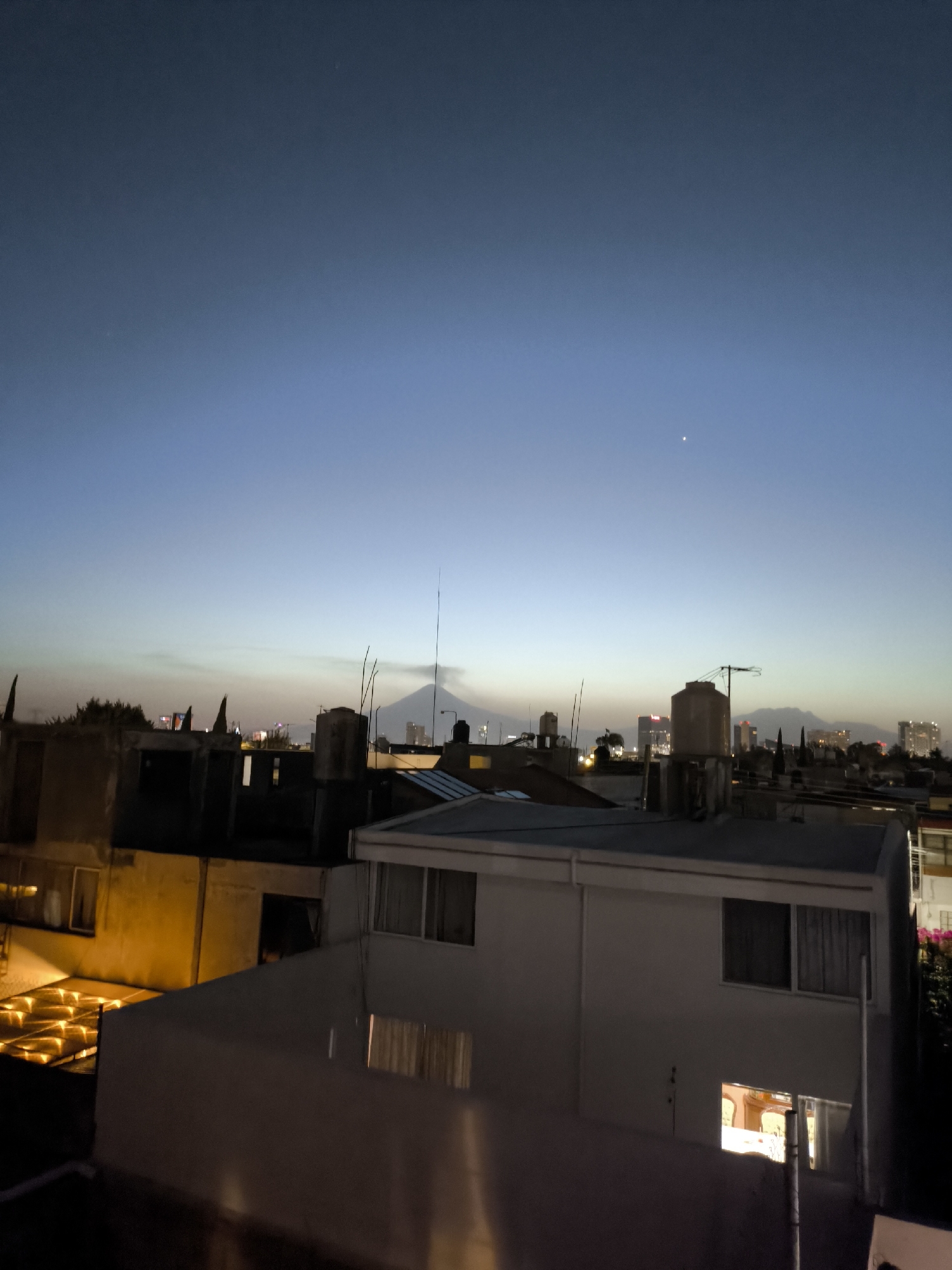 The image shows a twilight cityscape in Puebla, Mexico. The scene is dominated by rooftops of buildings, some lit internally, with a view towards the horizon. A mountain range, including what appears to be Popocatépetl volcano, is visible in the distance. Several antennas and water tanks punctuate the skyline atop the buildings. The sky is a gradient of blue and grey, with a single bright star or planet visible.