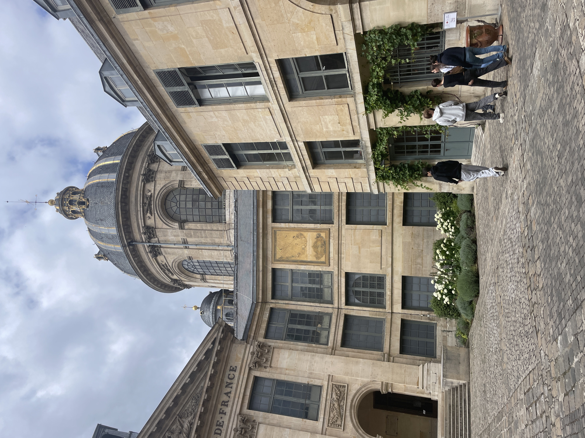 The image depicts the inner courtyard of the Institut de France in Paris, France, a grand architectural complex with historical significance. The central focus is a magnificent domed building, likely the chapel of the Institut, with a dark gray roof and ornate gold detailing. The building's facade is constructed of pale stone, featuring large arched windows and classical architectural elements.
To the left, another substantial stone building displays the inscription "DE FRANCE" above a pediment adorned with sculpted figures. This structure also has tall, dark-paned windows and a grand arched entryway with steps leading up to it.
To the right, a lighter stone building with a more modern yet complementary architectural style houses several windows, some of which are open. This building is partially covered in ivy, adding a touch of natural green to the scene.
The courtyard itself is paved with cobblestones, creating a textured and historic ground surface. In the middle ground, a well-maintained garden area features a profusion of white flowers and green shrubbery.
Several people are present in the courtyard. Four individuals are grouped together near the right-hand building, engaged in conversation. One person is walking away from the camera towards the left, while another is walking towards the group.
The sky above is overcast with gray clouds, suggesting a temperate climate. The overall impression is one of historical grandeur, intellectual pursuit, and a serene urban courtyard environment within the heart of Paris.