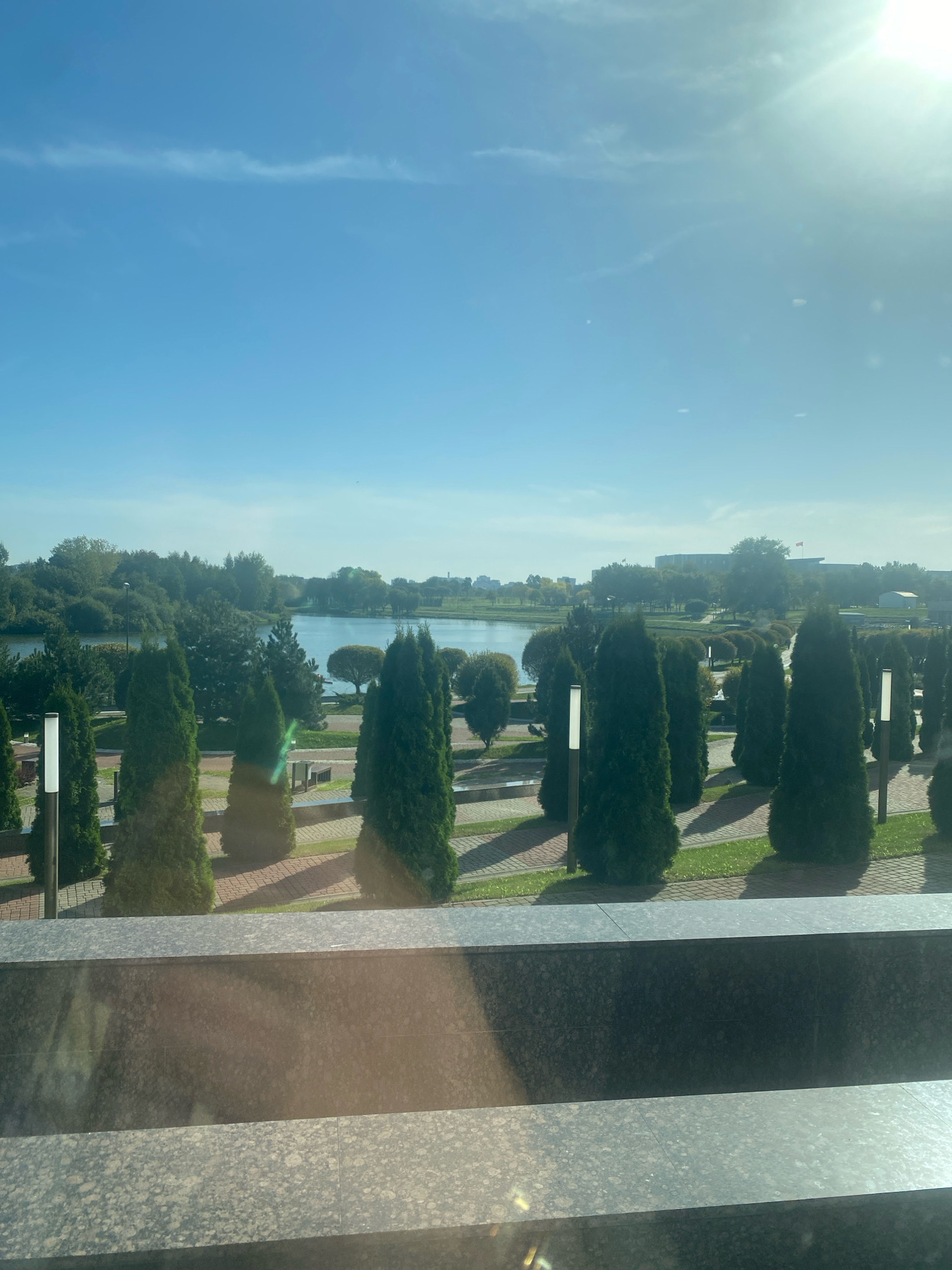 The image shows a view of the Svislach River and the Island of Tears memorial complex in Minsk, Belarus, on a sunny day. In the foreground, there are several rows of manicured evergreen trees, likely cypresses, planted in a formal garden setting. Modern outdoor lamps are placed between the trees. The ground is paved with brick and grass. A body of water, the Svislach River, is visible in the middle ground, with trees lining its banks. In the background, there are buildings and more greenery, typical of a city park or waterfront area. The sun is bright, casting shadows and creating lens flare in the upper right corner of the image. The overall impression is one of a well-maintained public space with a serene natural backdrop.