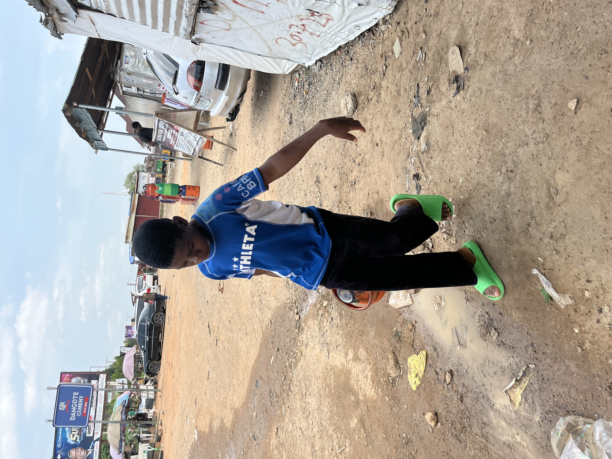 A young boy walks forward on a dirt road in Abuja, Nigeria, carrying an orange bucket in his right hand. He wears a blue and white athletic shirt and black pants, along with bright green slide sandals. The road is wet and muddy in places, with scattered debris. In the background, a large billboard for Dangote Cement is visible, along with various structures, vehicles, and people, suggesting a busy marketplace or roadside area. The sky is overcast.