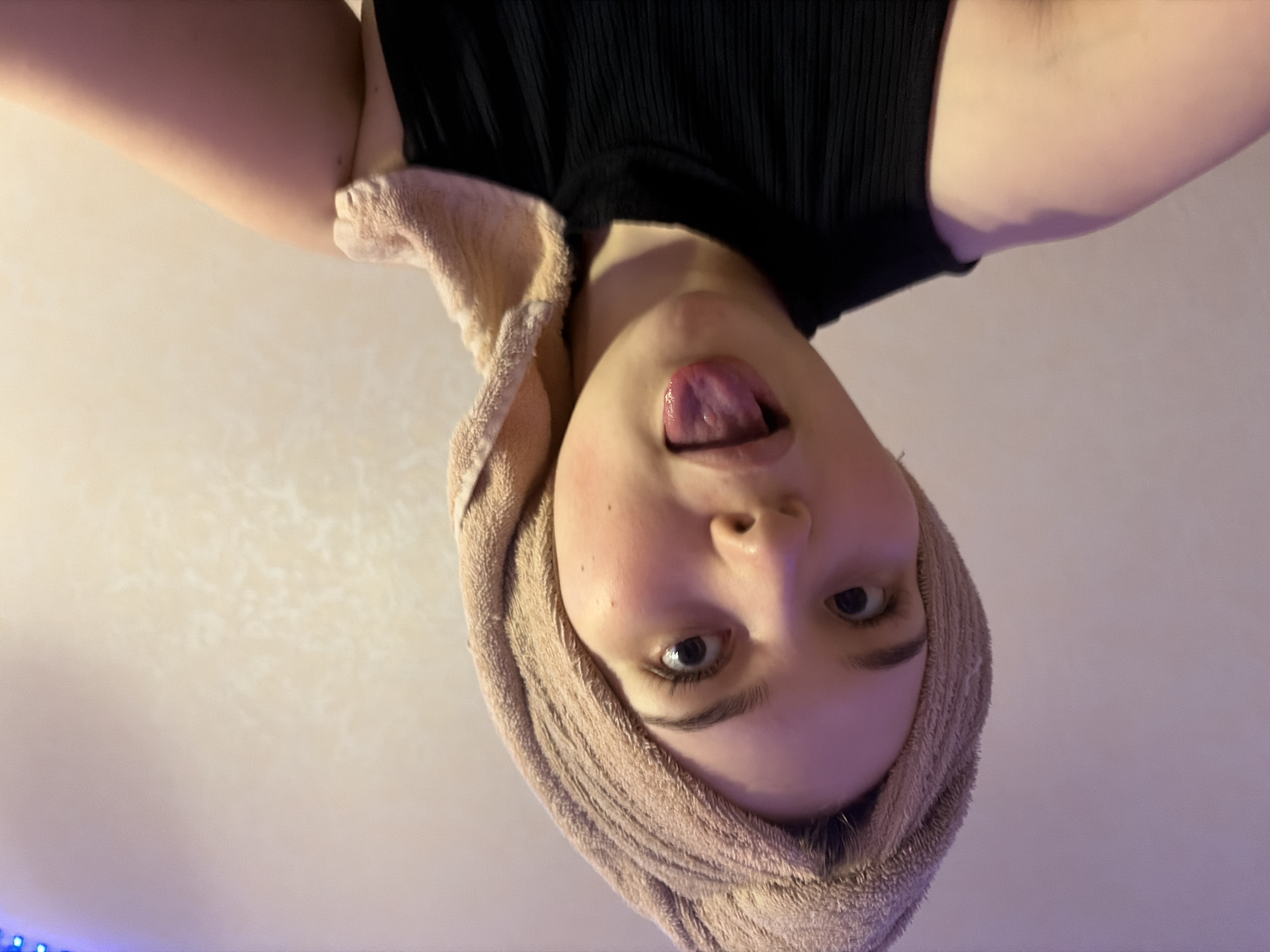 A Caucasian woman with a fair complexion is looking directly at the camera from an upside-down perspective. She is wearing a black, ribbed tank top, and a beige towel is wrapped around her head, framing her face. Her mouth is slightly open, with her tongue partially extended.
The background appears to be a light-colored wall or ceiling with subtle variations in texture. A string of blue lights is visible in the lower-left corner. The photo appears to have been taken in an indoor environment. There is no specific information that indicates the location is Chaikovsky, Russia.