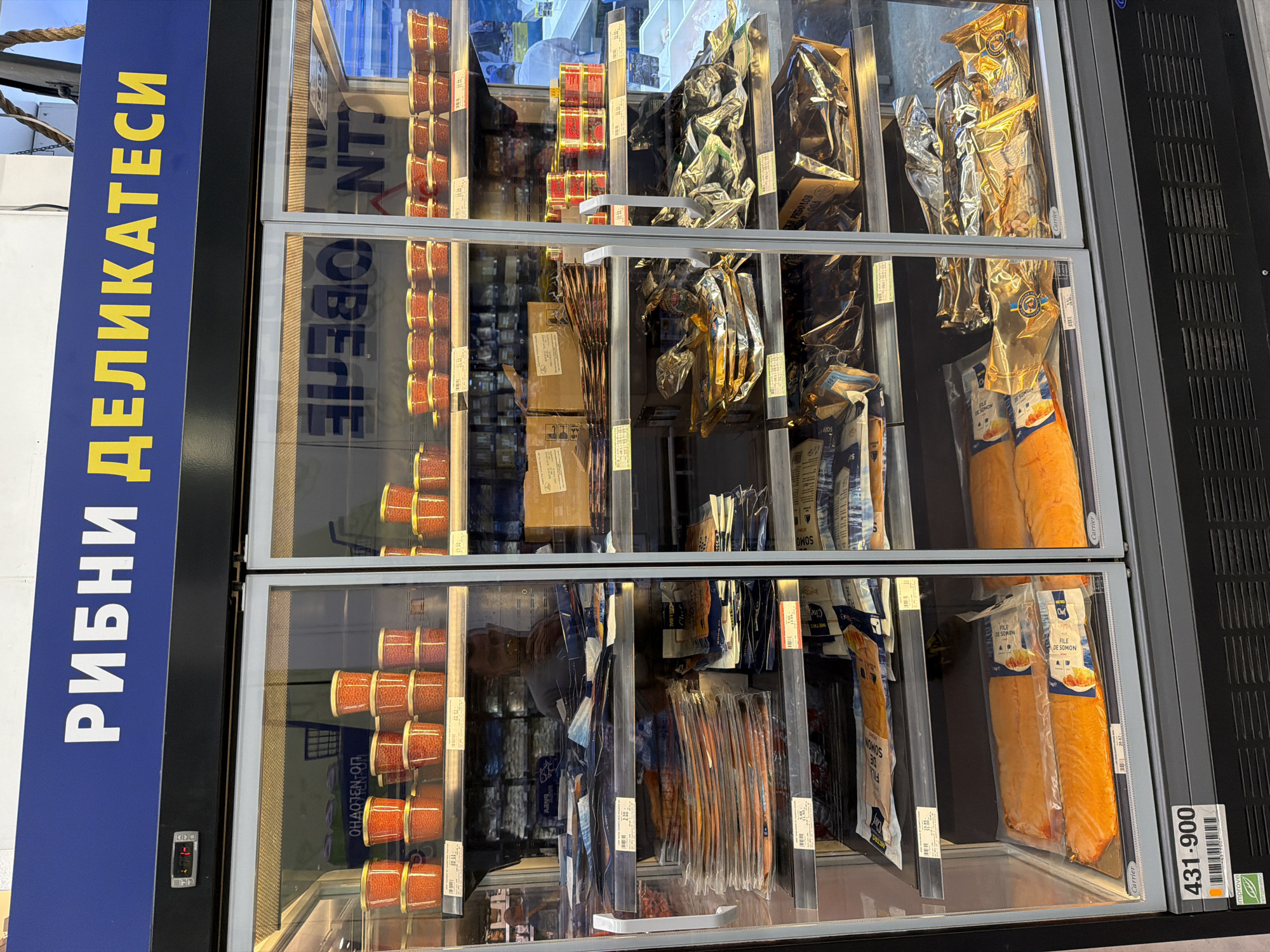 This image shows a refrigerated display case filled with various seafood products. The sign on the left side of the case is written in Cyrillic, specifically Bulgarian, and translates to "Fish Delicatessen". The display case has three shelves with a variety of items. The left side of each shelf is stacked with small, round containers, likely containing caviar or fish roe. The middle section of each shelf contains individually packaged seafood items, with various shapes and sizes. The right side of each shelf contains longer, flat packages of what appears to be salmon fillets. The price tag "431-900" is displayed on the right side of the bottom shelf. The bottom two shelves contain fish filets. In the city of Burgas, Bulgaria.