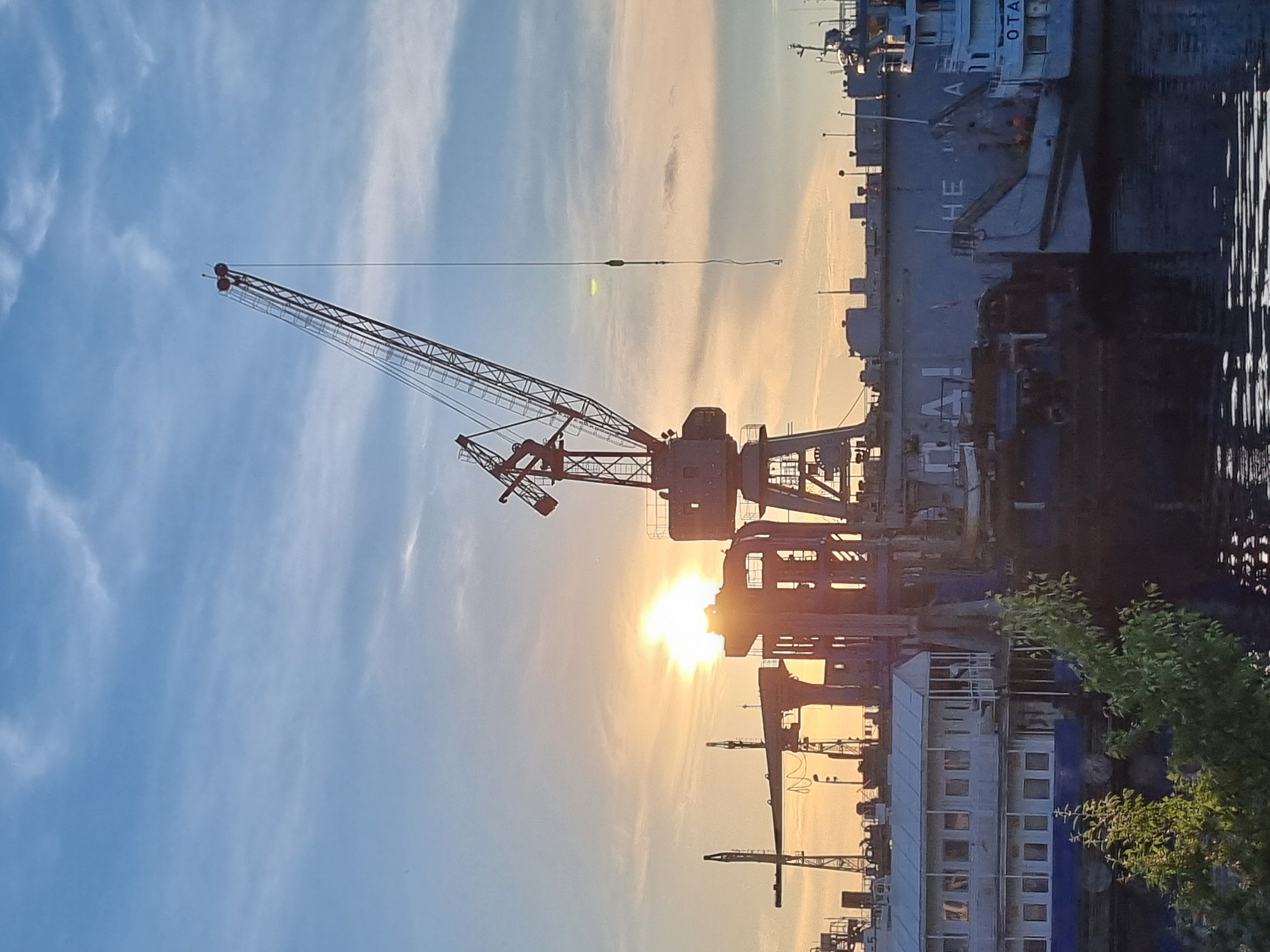 An industrial waterfront scene is captured at sunset in Chaikovsky, Russia. Dominating the frame are several large cranes, silhouetted against a pale orange and blue sky filled with wispy clouds. The sun, a bright orb, is partially obscured by one of the structures, casting a warm glow over the scene. In the foreground, a body of dark water reflects the sky and industrial elements.
To the left, a multi-story building with white siding and blue trim is docked. Behind it, and stretching across much of the mid-ground, is a large, dark-colored barge or ship. Visible lettering on its side reads "HE H A", part of a larger word. Another ship, predominantly white, is docked to the right of the barge, with "OTA" visible on its hull.
Several smaller structures and cranes are visible further in the background. Green foliage is present in the lower left corner, suggesting the presence of trees or bushes near the water's edge. The overall impression is one of a working port or shipyard at the close of day, with industrial machinery and vessels set against a dramatic sunset.