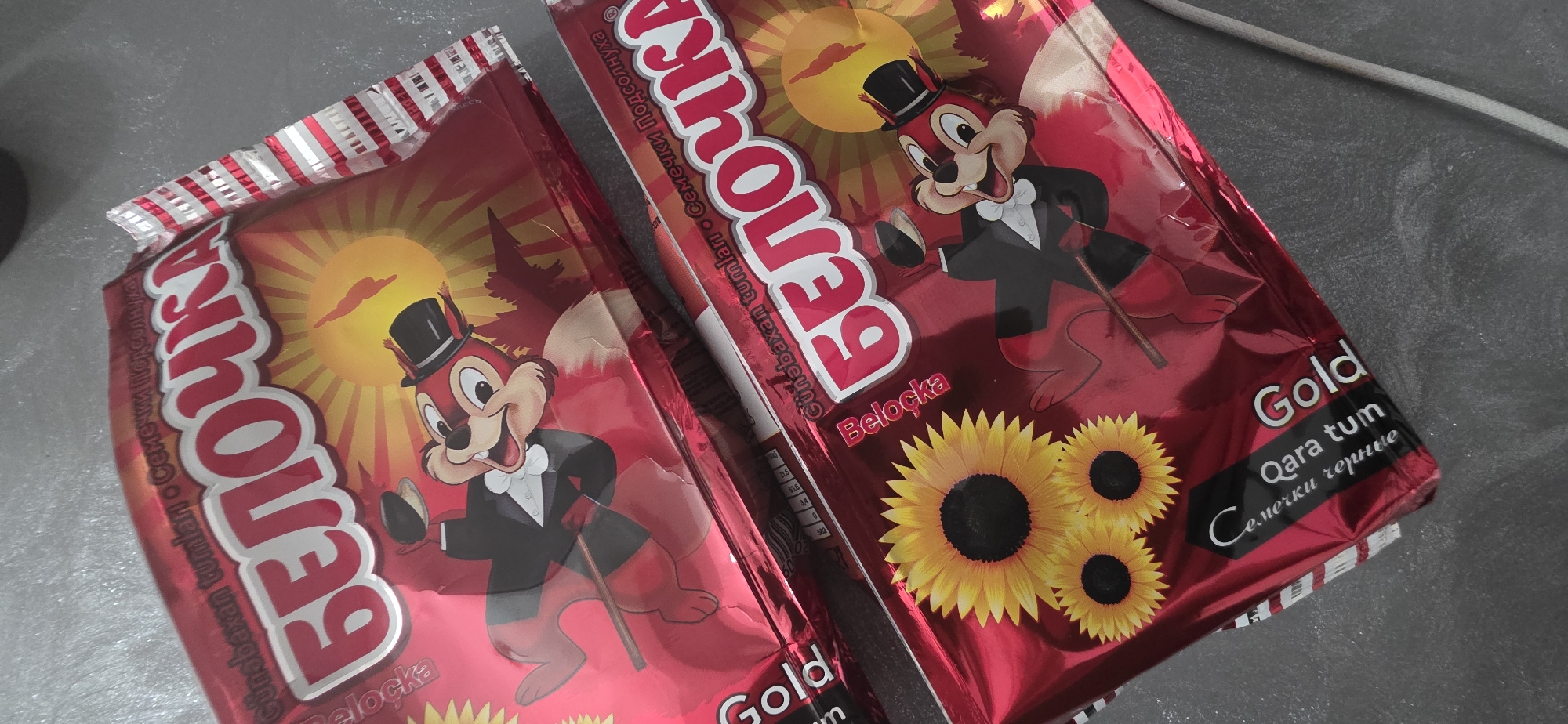 The image shows two packages of "Belochka" brand sunflower seeds. The packaging is predominantly red with gold accents and features a cartoon character of a squirrel dressed in a tuxedo and top hat. Sunflowers are also depicted on the packaging. The packages are lying on a dark grey, textured surface, possibly a tabletop. The lighting suggests an indoor environment. There are no people visible in the image. The scene appears to be a still life arrangement of the product. No specific location in Sumgayit, Azerbaijan, can be identified from the image itself.