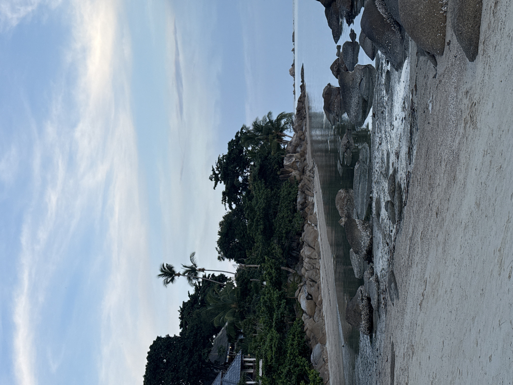 A tropical beach scene in Ko Samui District, Thailand, captures the early morning or late evening atmosphere. The foreground features a sandy beach with scattered rocks, some of which are partially submerged in the calm, reflective water. Lush green vegetation and palm trees line the shore, interspersed with large boulders. A building with a thatched roof and open-air verandas is nestled among the trees on the left side of the frame. The sky above is a soft blue with wispy white clouds. The overall impression is one of tranquility and natural beauty.