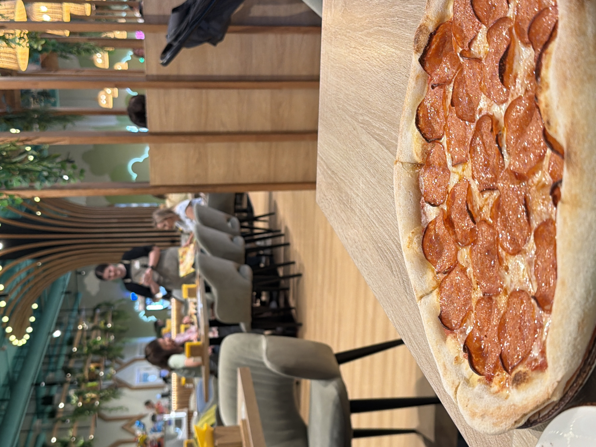 A close-up shot focuses on a pepperoni pizza, its crust golden brown and topped with generously spaced slices of pepperoni glistening under warm light, resting on a light-colored wooden table. In the blurred background of what appears to be a restaurant in Kazan, Russia, several patrons are visible seated at tables, engaged in conversation or dining. A server wearing an apron stands in the mid-ground, holding a notepad. The interior design features warm wood accents, hanging greenery, and decorative lighting, suggesting a modern and inviting atmosphere. The scene captures a casual dining experience.