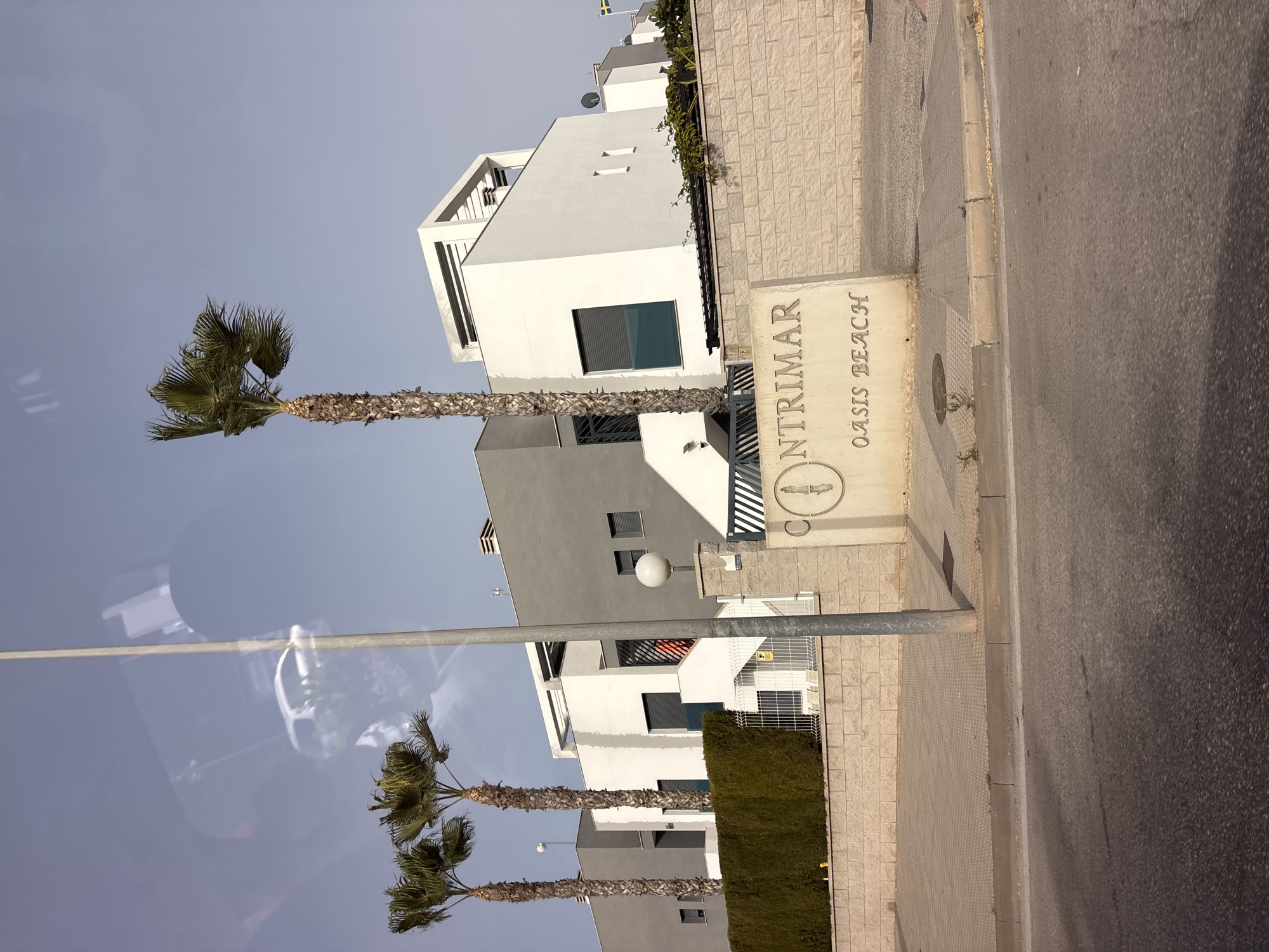 The image shows a street view in Orihuela, Spain, featuring modern residential buildings and palm trees. The buildings are white and gray, with clean lines and geometric designs. One building has a prominent white structure on top. In front of the buildings is a low, textured brick wall topped with a hedge and a metal fence. A large sign is partially visible, with the words "CONTRIMAR" and "OASIS BEACH" inscribed on it, along with a circular emblem containing a depiction of a knife or sword. Two tall palm trees are visible, with one on the right being particularly tall and slender. A light pole stands in the foreground, casting a shadow on the pavement. The sky is clear and pale blue. A reflection of a white car and a tall light pole can be seen in the upper left portion of the image, suggesting it was taken through a window. A small Swedish flag is visible on a distant building. The street in the foreground is asphalt, with a concrete curb and patterned sidewalk.