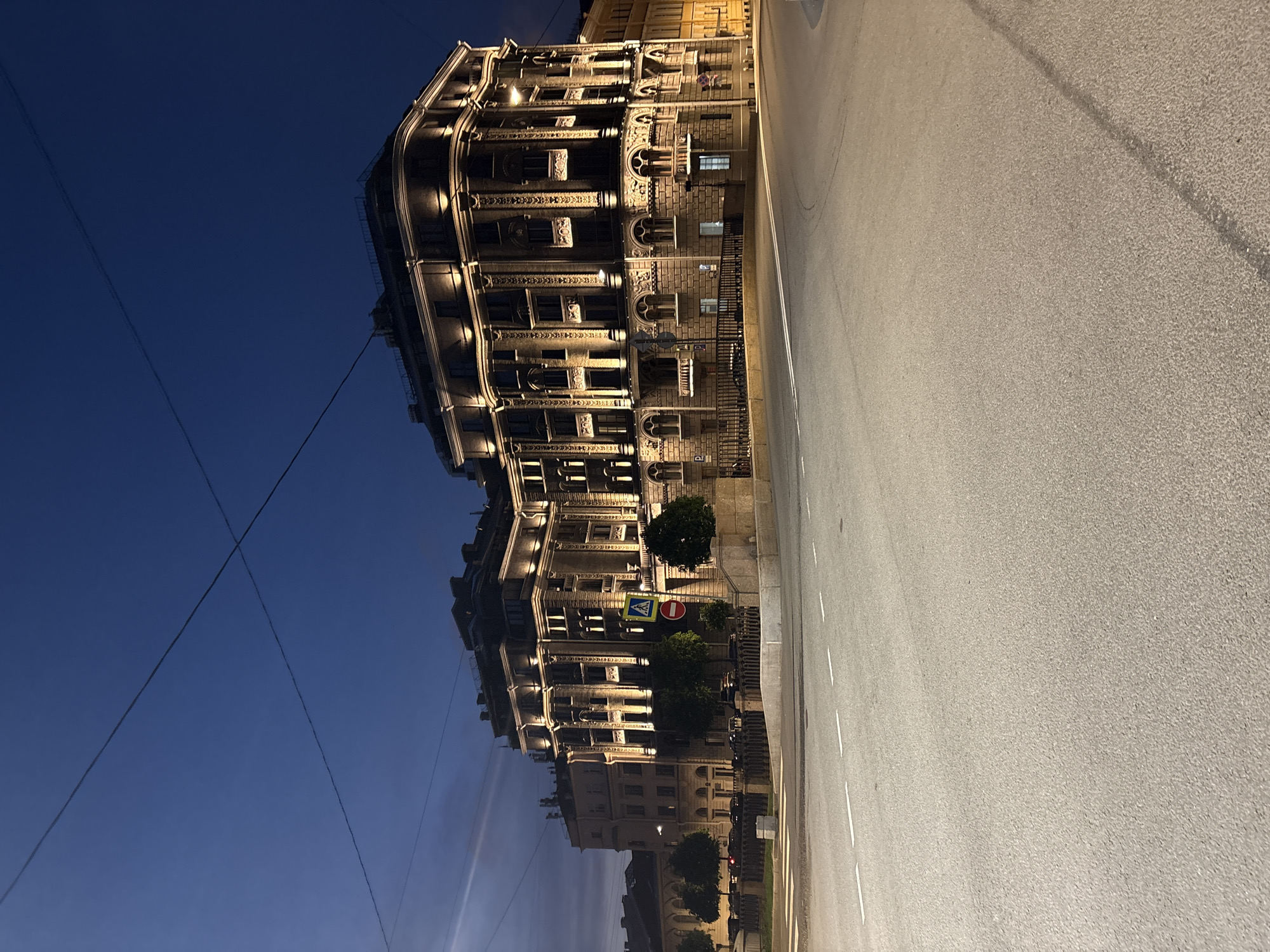 The image shows a street view in Saint Petersburg, Russia, at night. A multi-story building with ornate architectural details is illuminated with warm light, contrasting against the dark blue sky. Power lines are visible in the sky. A wide, paved street stretches to the right of the building, with painted white lines marking lanes. Trees and street signs are visible near the building.