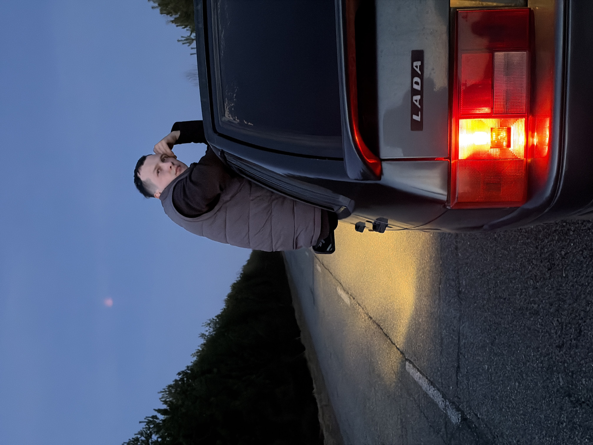 An outdoor scene captured at dusk or early night in Kharnavy, Russia, shows a man leaning out of the passenger side window of a dark-colored car. The man, with short dark hair, wears a dark long-sleeved shirt and a grey puffer vest, looking upwards or towards the camera with a neutral, slightly pensive expression, his right hand resting near his temple.
The car, which appears to be stationary, is positioned on the side of an asphalt road. The right rear section of the car is prominent, featuring a brightly illuminated taillight that casts a distinct yellow glow onto the adjacent road surface. Vertically aligned next to the taillight, the brand name "LADA" is clearly visible on the car's trunk lid. A faint white line marks the edge of the road. In the background, the sky transitions from dark blue overhead to darker tones near the horizon, with the silhouette of trees or bushes lining the roadside. The low light conditions are accentuated by the car's activated taillight, suggesting twilight hours.