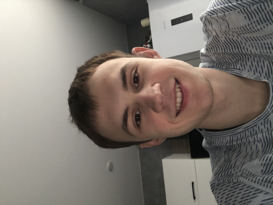 The image displays a head-and-shoulders selfie of a young adult male in an indoor domestic setting. The specific location within Krasnodar, Russia, is not ascertainable from the visual information.
**Person:** The subject is a light-skinned male with short, dark brown hair and brown eyes. He is looking directly at the camera, exhibiting a wide smile with his teeth visible. He wears a light-colored t-shirt with a repeating pattern of thin, dark, wavy horizontal lines.
**Environment and Objects:** The background suggests a kitchen or dining area. To the left, a portion of a white cabinet or furniture unit with a black horizontal handle is visible, set against a dark grey textured wall. To the right, a white refrigerator with a dark vertical control panel is present. Above the refrigerator, a small shelf holds what appears to be a white cylindrical object, possibly a candle. The ceiling is white and features a single recessed light fixture in the upper left quadrant of the frame. The lighting is uniform and soft, consistent with an indoor environment.
**Actions/Interactions:** The male subject is actively smiling, suggesting a positive disposition or engagement with the camera. No other individuals or interactions are depicted.