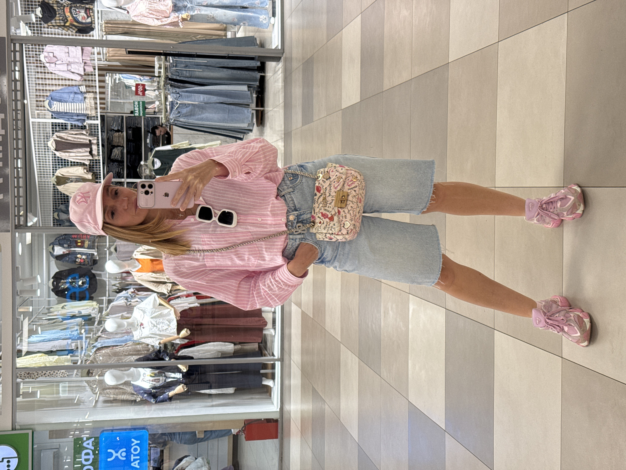 A woman is captured in a mirror selfie within what appears to be a clothing or department store, likely in Saint Petersburg, Russia given the context and visible text. She is standing upright, holding a pink iPhone with a light-colored case to take the picture, concealing her face partially.
She is dressed in a casual, summer-appropriate outfit, featuring a pink and white striped long-sleeved button-down shirt, light-wash denim shorts with frayed hems, and a light pink baseball cap with a white "NY" logo. A pair of white-framed sunglasses is tucked into her shirt collar. A small, light-colored shoulder bag with a floral and text pattern, featuring a chain strap, hangs across her body. Her footwear consists of chunky pink and white athletic sneakers.
The background, visible in the mirror's reflection, reveals numerous clothing racks displaying various garments like jeans, shirts, and jackets. Several white mannequins are also visible, dressed in clothing. The floor is tiled in an alternating pattern of light and dark gray or beige stripes. Reflections of overhead lights are present.
Visible text in the background, on various signs and shop displays, includes Russian words such as "РАБОТА" (work), "ОДЕЖДА" (clothes), and fragments like "СКИДКА" (discount) and "АТOY" which further indicate a Russian-speaking location. The woman's expression is neutral as she poses for the photo. The setting suggests daytime, during business hours.