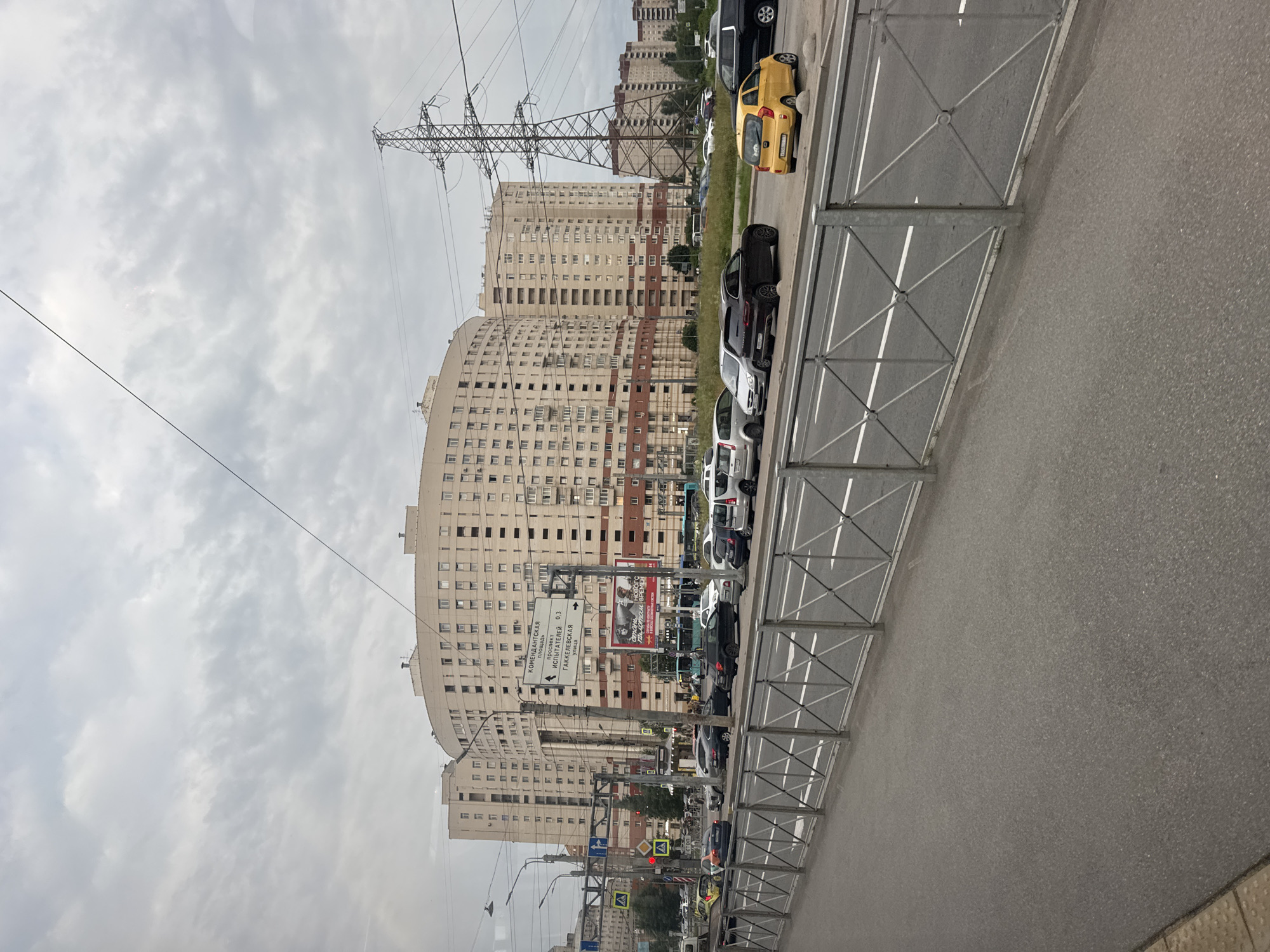 This image captures a street scene in Saint Petersburg, Russia, under an overcast sky. A large, curved, light-colored apartment building dominates the background, featuring multiple windows and balconies. To the right, a tall, lattice-style electrical pylon stands prominently against the sky.
In the midground, a wide, multi-lane road is visible, with several cars parked and in motion. A yellow car is parked on the right side of the road. Traffic lights and street signs are present, including a large directional sign with Cyrillic text indicating "Komendantskaya Square" and distances to "Prospekt Ispolniteley" and "Gakkelevskaya Ulitsa." A large billboard featuring images of people is also visible.
The foreground shows a paved walkway separated from the road by a metal fence with a diamond pattern. The ground is asphalt, with some grass and small bushes growing along the edge of the road. The overall impression is of a typical urban environment in Russia.